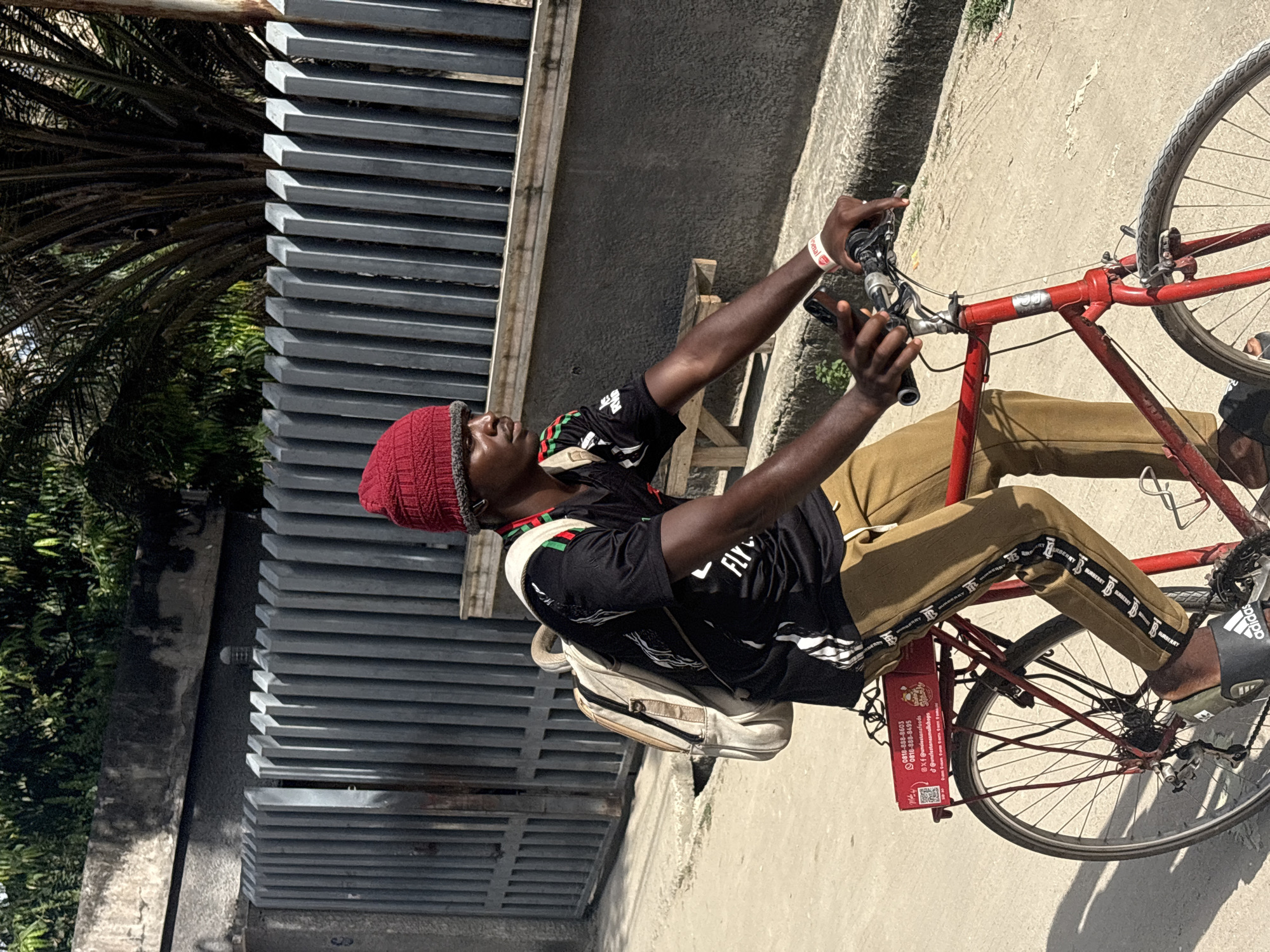 A man with dark skin, wearing a red knitted hat, a black t-shirt, and khaki pants with Burberry branding, is sitting on a red bicycle in Igboefon, Nigeria. He has a backpack on and is holding a smartphone. The bicycle has a red box attached to the back. The background includes a gray metal structure, trees, and a concrete sidewalk. The photo is taken at an angle.