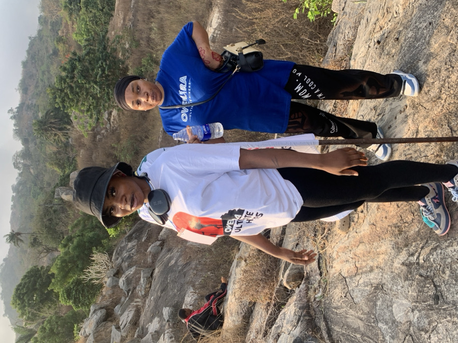 Two women stand on a rocky, mountainous landscape. The foreground figure wears a white t-shirt, black hat, and headphones around her neck. Behind her, the other woman wears a blue t-shirt, black pants with "I'M A COOL MOM" on them, and a black head covering. A water bottle is in her hand. A metal rod is visible between them. Rocky terrain and sparse vegetation cover the mountain; a hazy view of the landscape stretches into the distance.