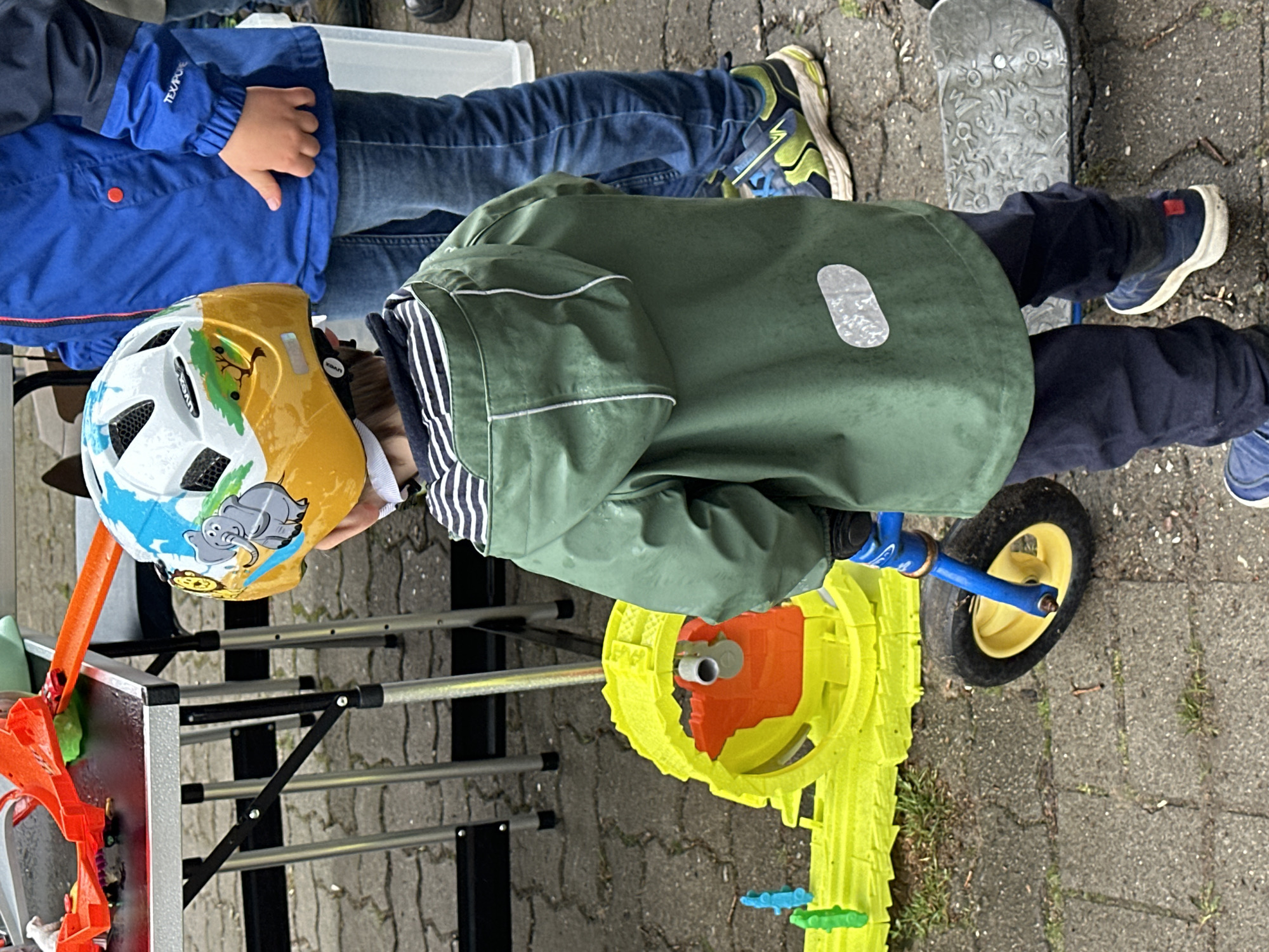 This image captures an outdoor scene in Deinste, Germany. The primary subject is a young child, viewed from behind, standing on a paved surface. The child wears a white, yellow, and blue Uvex helmet featuring grey elephants and green tree motifs. A dark green raincoat with white reflective piping and a rectangular white reflective patch on the lower back covers a black and white striped long-sleeved shirt. Dark blue trousers and blue sneakers with white soles and a red heel accent are worn. The child is holding the handlebars of a blue and yellow balance bike, with its yellow front wheel resting on the pavement.
To the right and slightly behind the foreground child, another individual, partially visible from the torso down, is also viewed from behind. This person wears a blue jacket with "TEX/PURE" visible on the sleeve, blue jeans, and blue, green, and black athletic shoes with prominent white soles. Their right hand is visible to their side.
In the background, to the left of the foreground child, a portable table with silver legs and a dark surface is set up. An orange toy race track, complete with ramps and a small red toy car, is arranged on the table. On the ground to the left, a large, bright yellow and orange plastic toy structure, possibly a segment of a play track or tunnel, is visible, with small blue and green plastic figures positioned nearby. To the immediate right of the foreground child's feet, a grey skateboard featuring an embossed pattern of flowers and stars rests on the ground.
The ground consists of interlocking grey concrete pavers, interspersed with small patches of dirt and sparse grass. The environment suggests an outdoor play area or event, under what appears to be overcast conditions.
