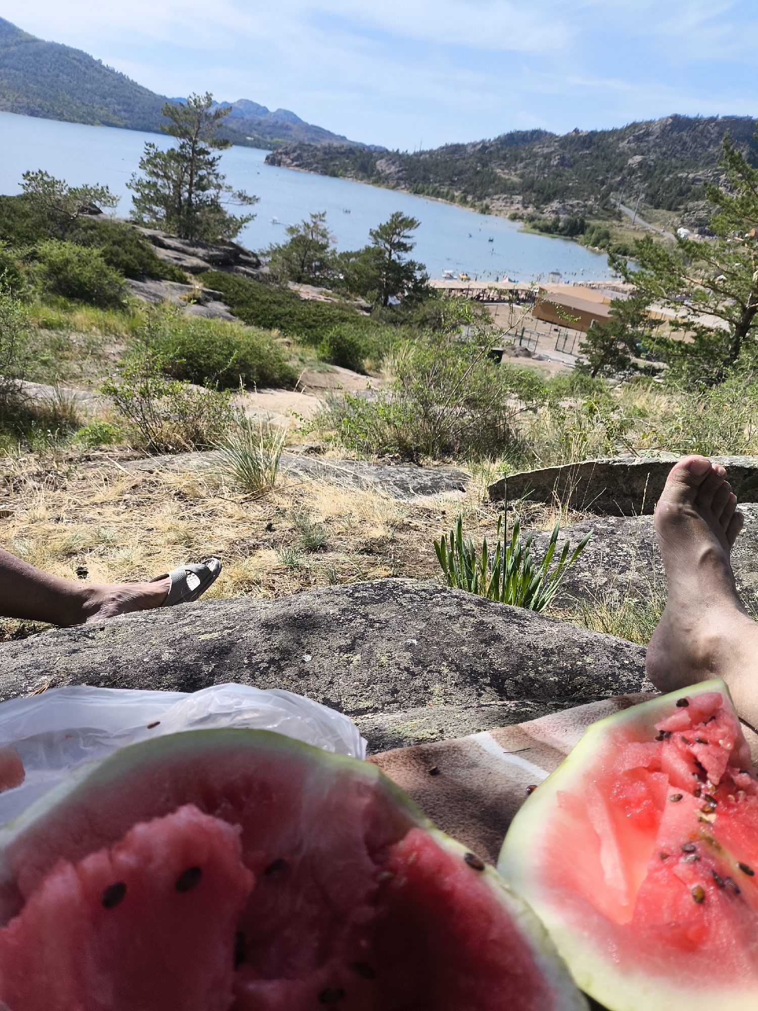The image depicts a scenic overlook near a body of water, likely Lake Sabyndykol in Bayanaul National Park, Kazakhstan. In the foreground, two partially visible human feet are resting on a rocky surface, suggesting a casual, outdoor setting. One foot is wearing a grey sandal. A partially eaten watermelon is spread out on a striped cloth in front of the feet, with a clear plastic bag nearby. The middle ground shows a slope covered in dry grass and sparse green bushes, leading down to a busy beach area with numerous people and structures. The background features a wide, calm lake bordered by rugged, tree-covered hills and mountains under a bright, partly cloudy sky. The overall impression is of a relaxing day spent enjoying nature and a picnic.
