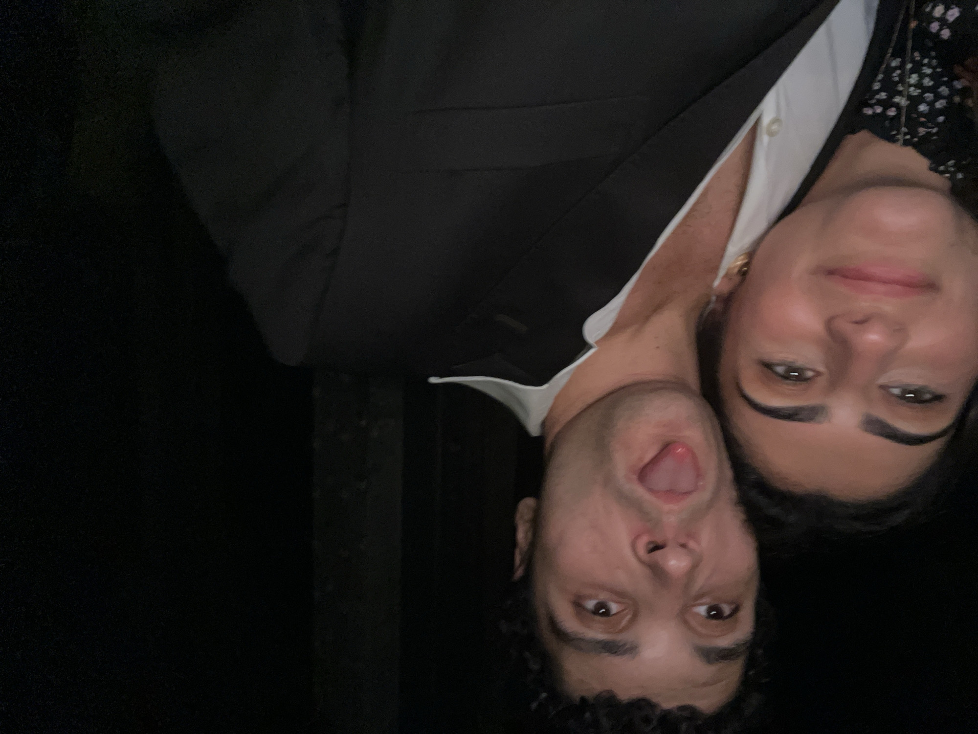 A close-up, waist-up selfie captures a young couple. The man, on the right, has curly dark hair and is sticking his tongue out at the camera. He wears a white collared shirt under a black suit jacket. The woman, on the left, has dark hair and is smiling and looking towards the camera. She wears a patterned dress and gold earrings. The background is dark and out of focus. The lighting suggests an indoor evening setting.