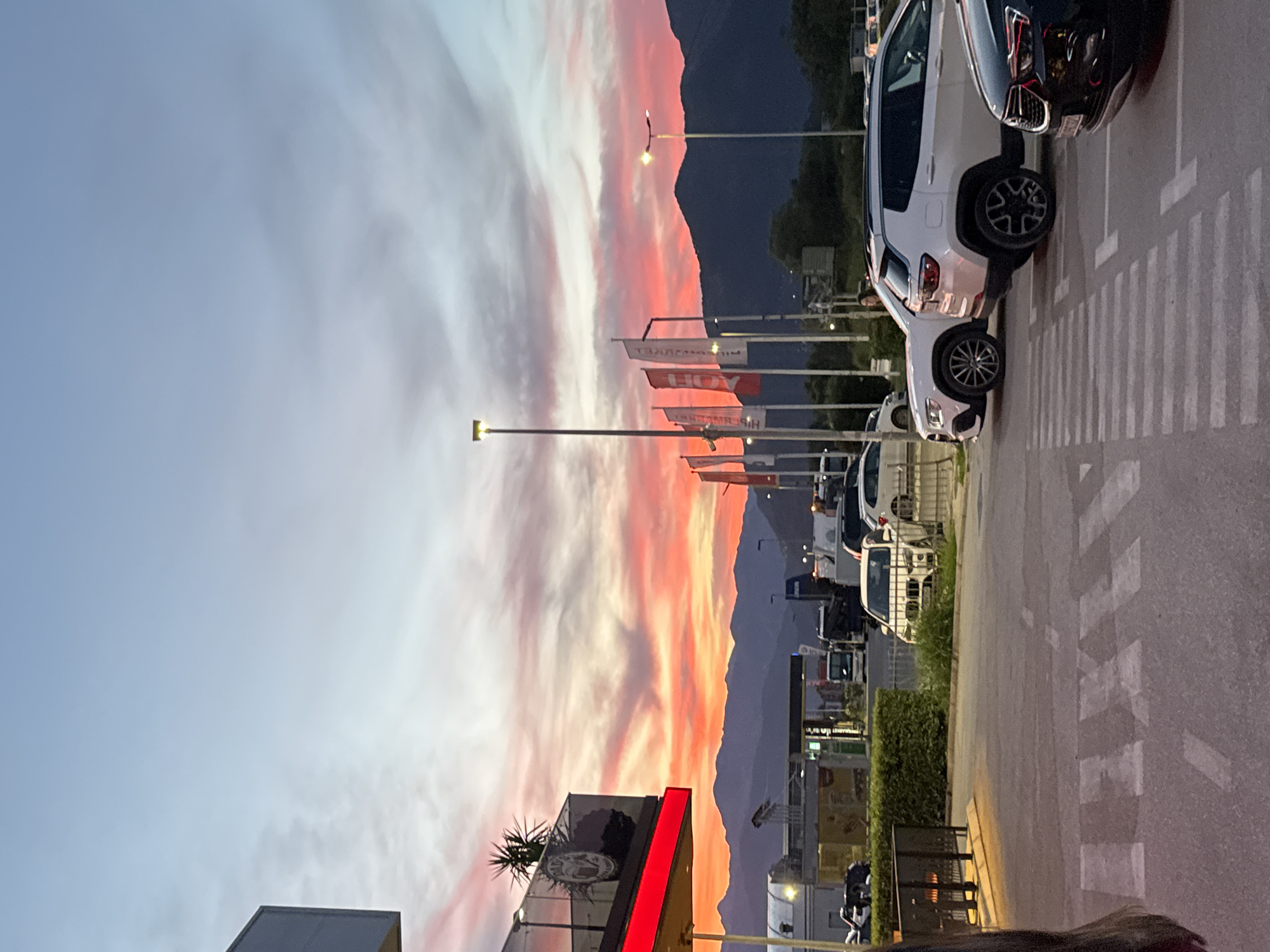 The image captures a sunset scene in Dub, Montenegro. The sky is a vibrant spectacle of pink, orange, and purple hues, with clouds streaking across the expanse. In the foreground, a parking lot is visible, with several cars parked, including white SUVs and a dark-colored sedan. A pedestrian crossing is marked on the asphalt. In the mid-ground, there are flags on poles, a gas station with a canopy, and various vehicles, including trucks. Beyond the immediate structures, a range of mountains forms a silhouette against the colorful sky. A building with a red awning and a plant on a balcony is visible on the left side of the frame. The overall impression is of a commercial area at dusk, with the natural beauty of the sunset providing a dramatic backdrop.
