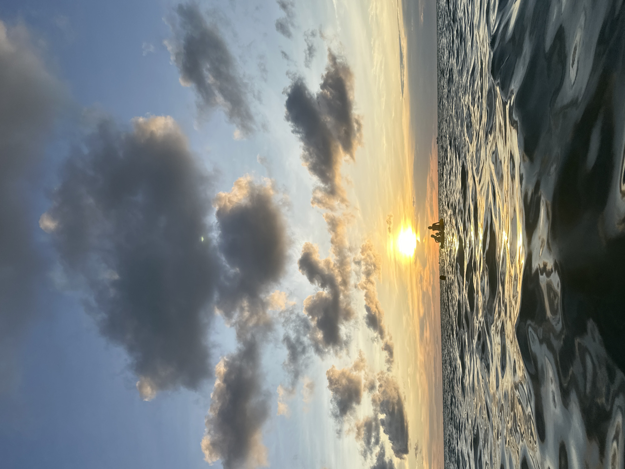 The image depicts a low-angle shot of the ocean at sunset, likely off the coast of Ko Phangan, Thailand. The foreground is dominated by rippling water with reflections of the sky. In the mid-ground, silhouetted figures are visible on what appear to be kayaks or small boats, positioned near the horizon where the sun is setting. The sky above is a blend of blue, orange, and gray clouds, with the sun casting a bright glow and reflecting on the water. The overall atmosphere is serene and picturesque, indicative of a tropical evening.