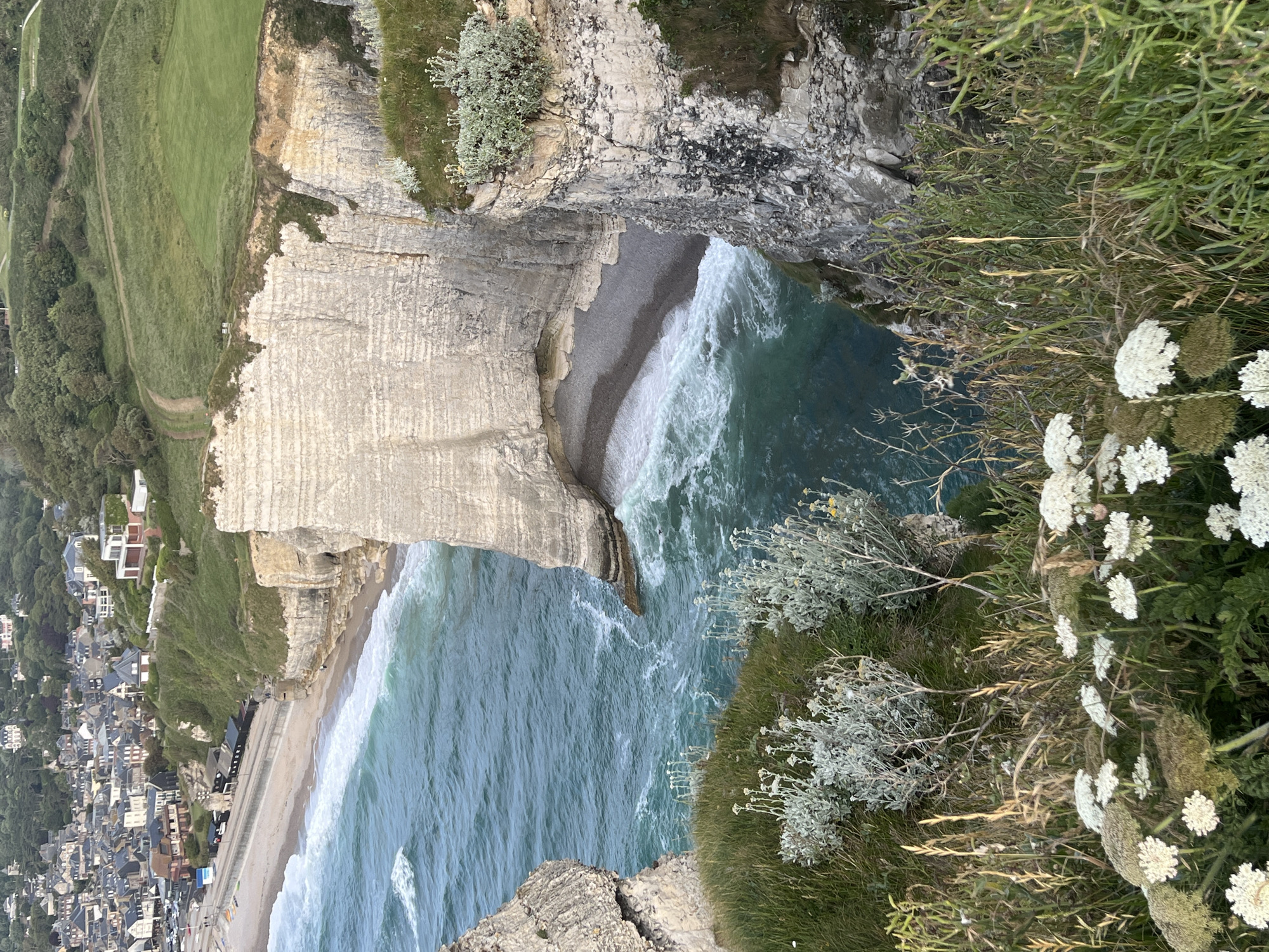 The image captures a scenic view of the cliffs and coastline in Étretat, France. In the foreground, wild white flowers and green vegetation grow on the cliff edge, overlooking the turquoise ocean. The water exhibits white waves crashing against the shore and the base of the cliffs. A small pebble beach is nestled within an archway of the imposing white chalk cliffs, which are a prominent natural landmark of Étretat. Further inland, a town with red-tiled roofs and houses is visible, situated on a hillside above a sandy beach lined with a promenade. The background consists of green hills and trees, with a dirt path winding through the landscape. The sky is overcast, suggesting potentially moody weather.