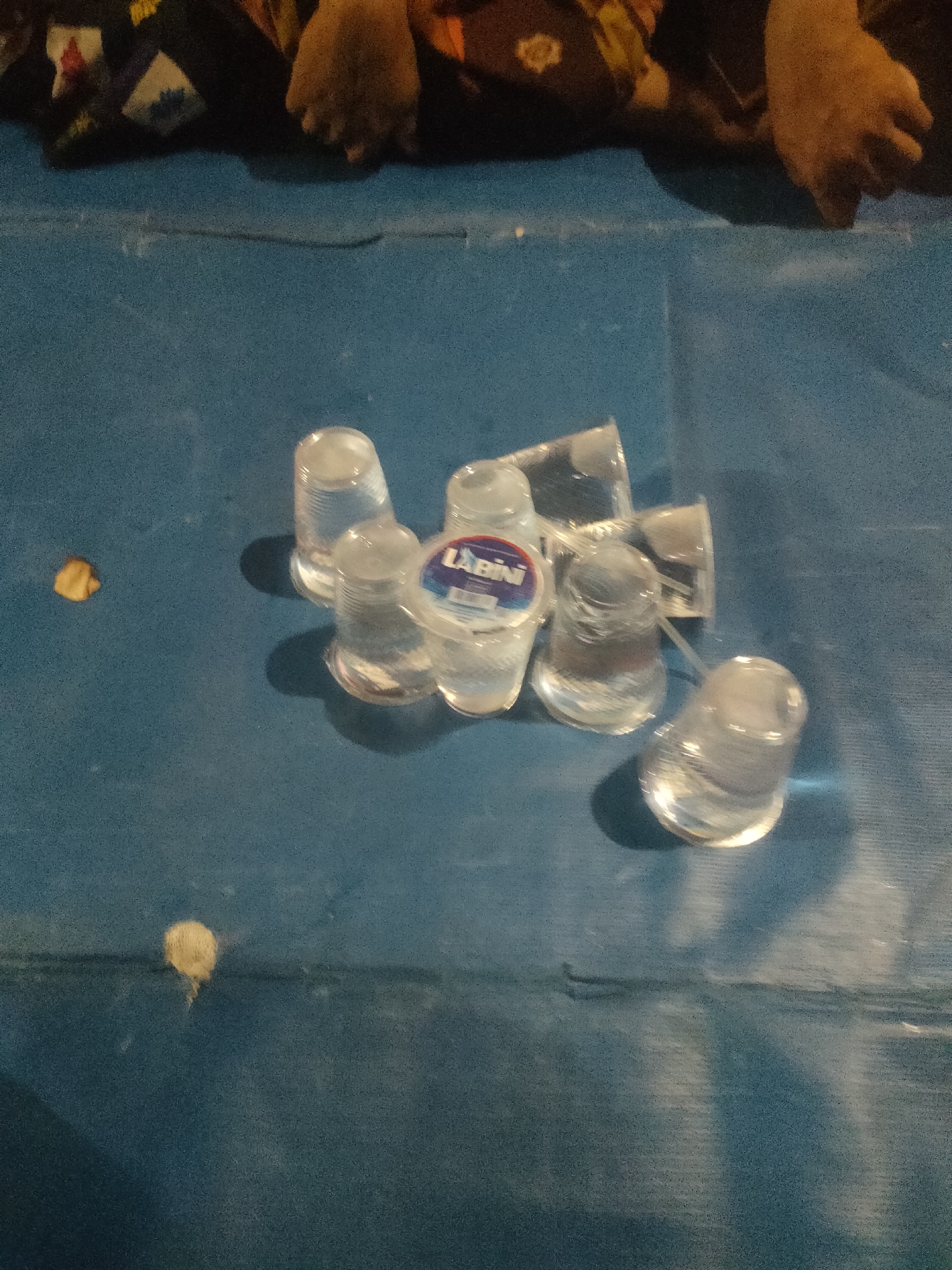 The image depicts a collection of clear plastic cups, some containing a clear liquid, scattered on a blue surface. One cup prominently displays a label with the text "ABINI". To the left of the cups, there is a small, yellowish-brown, irregular object. In the upper portion of the image, out of focus, are parts of human legs covered by dark fabric with some patterned detailing. The overall environment appears to be indoors or outdoors on a flat surface, possibly a table or the ground, under artificial lighting, casting shadows. The image does not contain any information that would allow for the specific location within the city of Surabaya, Indonesia.