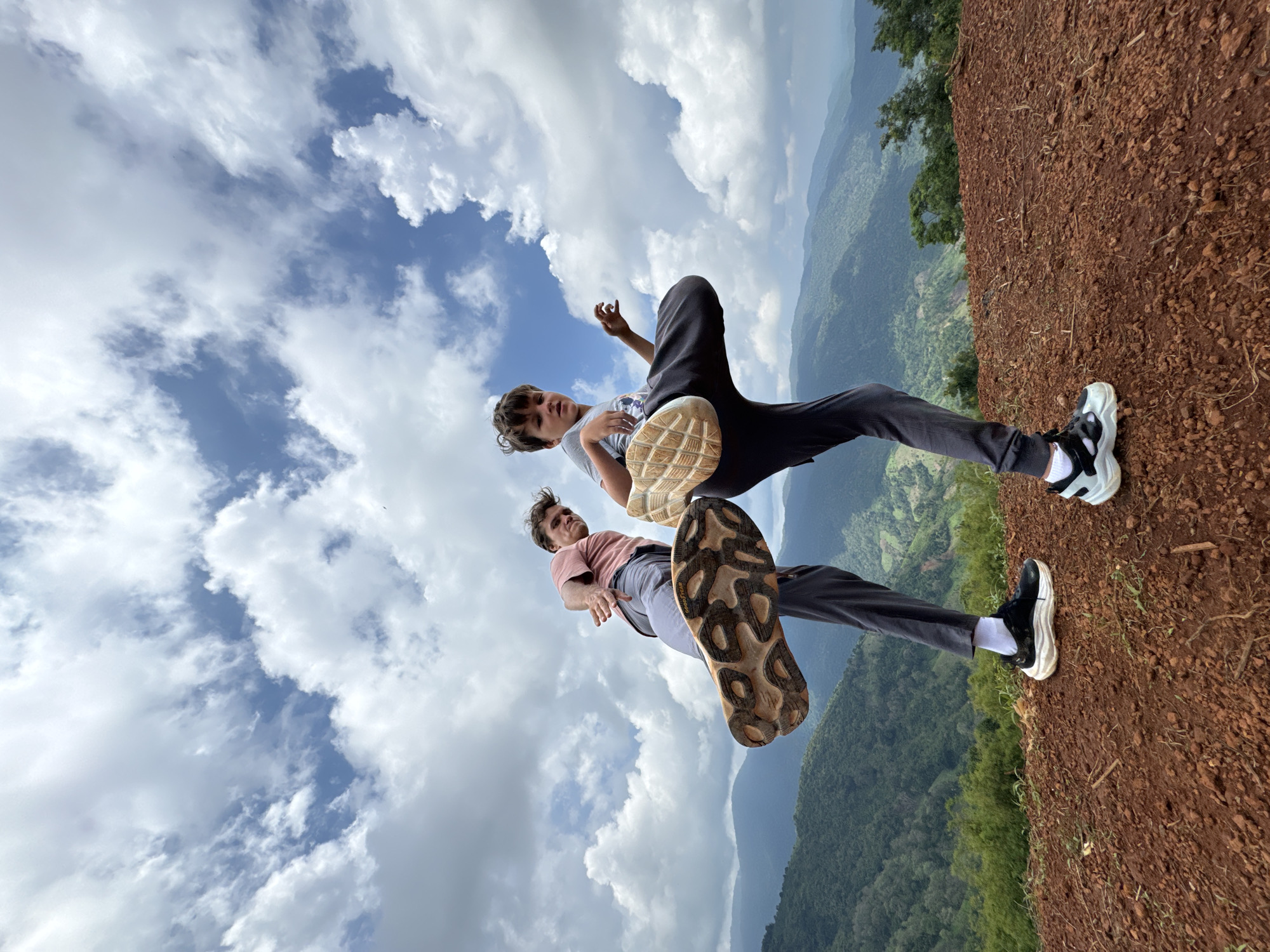Two young males are positioned on a reddish-brown dirt embankment, with one in front of the other, both facing away from the camera and kicking their legs outwards. The background consists of a lush, green, mountainous landscape under a cloudy blue sky. The person in the foreground, on the left, wears a pink t-shirt, grey pants, white socks, and athletic shoes with distinctive brown and black soles. The person behind and to the right wears a grey t-shirt, dark pants, and black and white athletic shoes. Their actions suggest a playful or martial arts-inspired pose. The overall setting appears to be a natural, outdoor location, likely a viewpoint overlooking a valley. This scene is not located in Nao Sẻ, Vietnam; it is a mountain landscape.