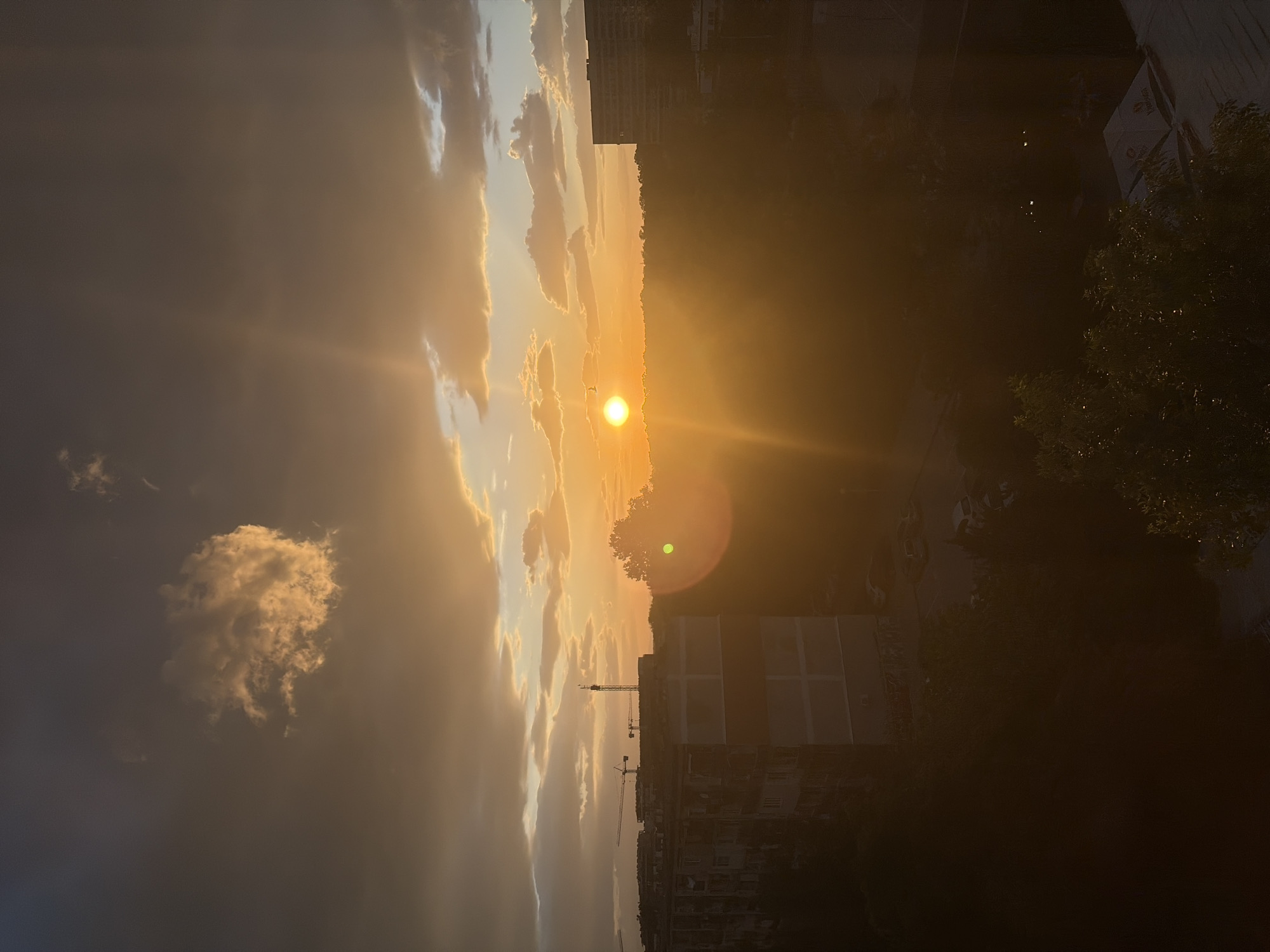 The image captures a vertical view of a sunset in what appears to be an urban area. On the left, the sky is filled with clouds ranging from dark grey to light yellow, illuminated by the setting sun. The sun is positioned near the horizon, emitting a bright, warm glow. To the right of the sun, buildings and trees are visible, silhouetted against the light. A construction crane can be seen in the distance. Given the urban setting and the presence of distinct buildings, this scene could possibly be located in Plovdiv, Bulgaria, although there is insufficient information to confirm this.
