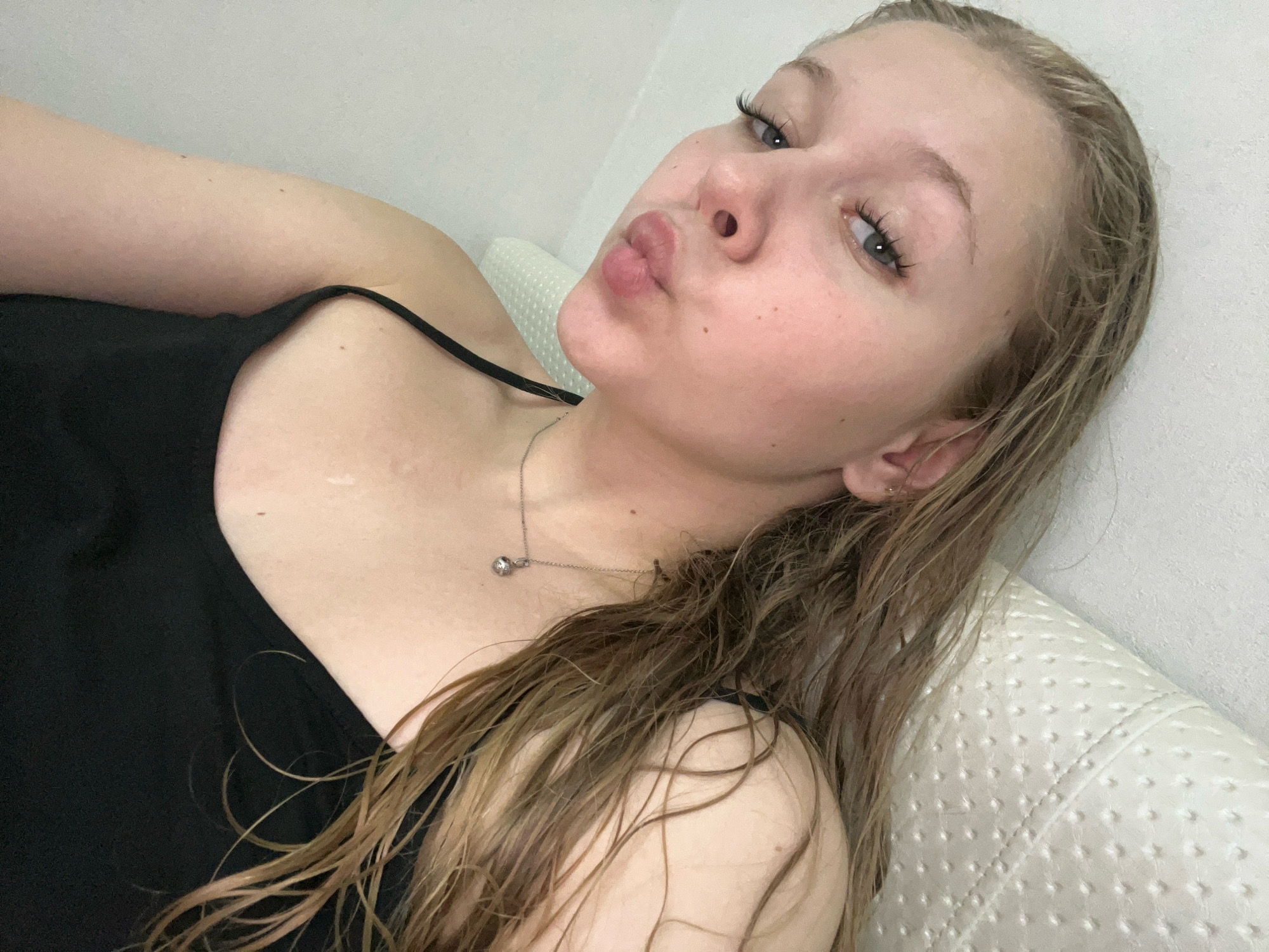 A young woman with blonde hair is lying on a textured, light-colored surface, possibly a pillow or headrest. She is wearing a black tank top and a delicate silver necklace with a small spherical pendant. Her hair is wet and falls around her shoulders and down her chest. She is looking towards the camera with her lips pursed in a kissing gesture. Her eyes are open, and she has noticeable eyelashes. The background is a plain, light-colored wall. There is no indication of the city of Perm, Russia, or any specific location within it.