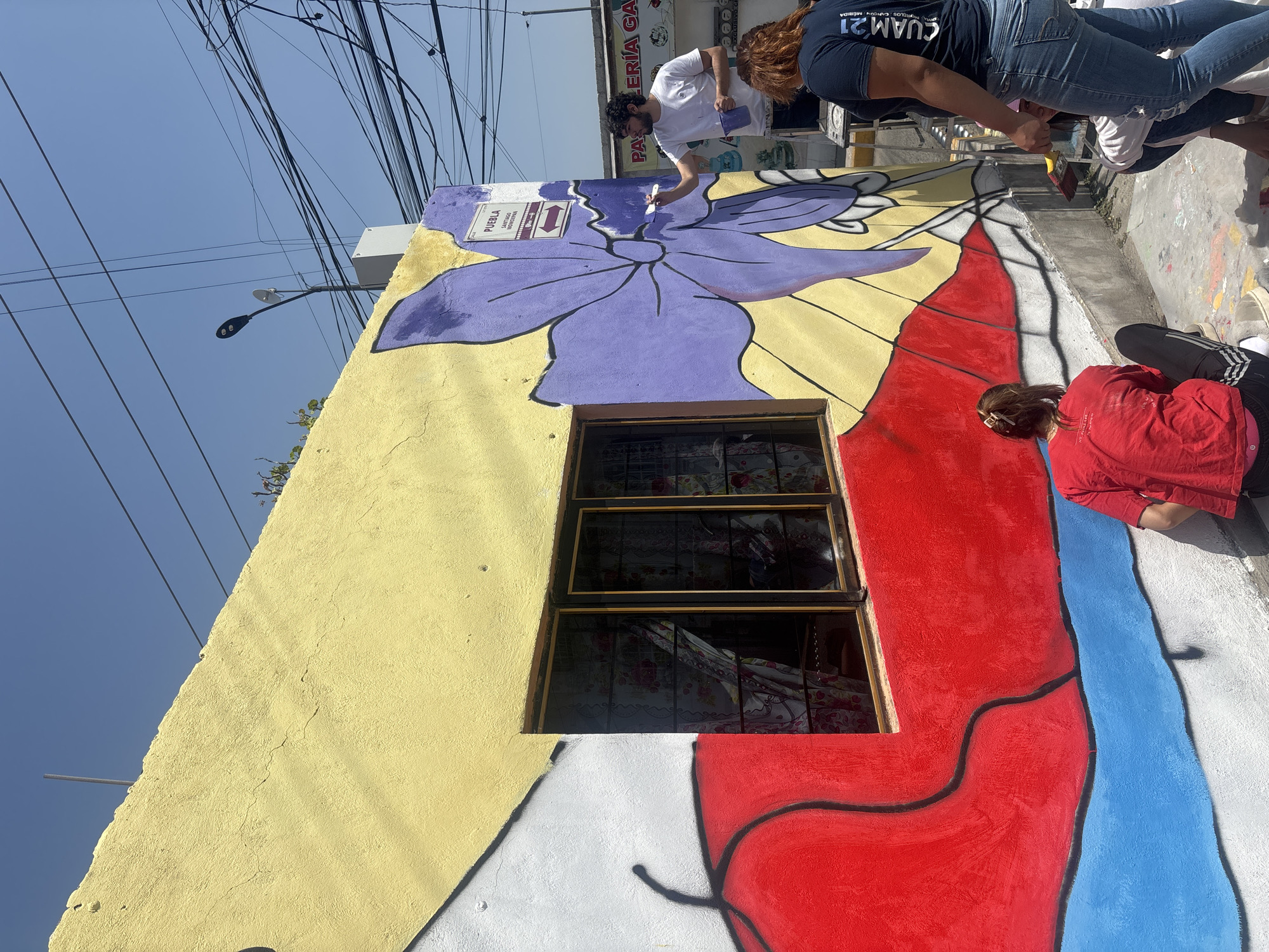 The image depicts an outdoor scene in Heroica Puebla de Zaragoza, Mexico, where several individuals are actively engaged in painting a colorful mural on the side of a building under a bright, clear blue sky.
**Subjects:** At least three people are visible, focused on the painting task. A man with dark, curly hair wearing a white t-shirt is painting a large purple flower on the upper part of the wall, holding a paintbrush and a small container of purple paint. Below him, a woman in a dark blue t-shirt and jeans is also painting, partially obscured. Further down, another person in a red long-sleeved shirt is seated or kneeling on the ground, working on a red section of the mural. The main subject is the building wall itself, featuring a textured yellow facade that serves as the canvas for the mural. A multi-paned window with dark glass and metal grates is incorporated into the mural's design. Overhead, numerous black electrical wires crisscross the blue sky, and a street light with two black lamps is visible.
**Activity or event taking place:** The primary activity is the creation or completion of a vibrant street mural. The people are actively applying paint to the wall, suggesting a community art project or an artistic endeavor to beautify the urban environment.
**Notable details:** The scene is illuminated by bright daylight, indicating a sunny day. The mural features a large, stylized purple flower with black outlines, set against the yellow wall. Below the window, the mural transitions into large, abstract shapes in red, white, and blue. The overall atmosphere appears collaborative and artistic.
**Visible text:** A white sign attached to the wall near the purple flower clearly reads "PUEBLA" with a directional arrow pointing left. Below "PUEBLA", some smaller text like "AYUNTAMIENTO" and "GOBIERNO" is partially visible. On the dark blue t-shirt of one of the painters, the text "GUAM'21" and "MERIDA" can be seen.