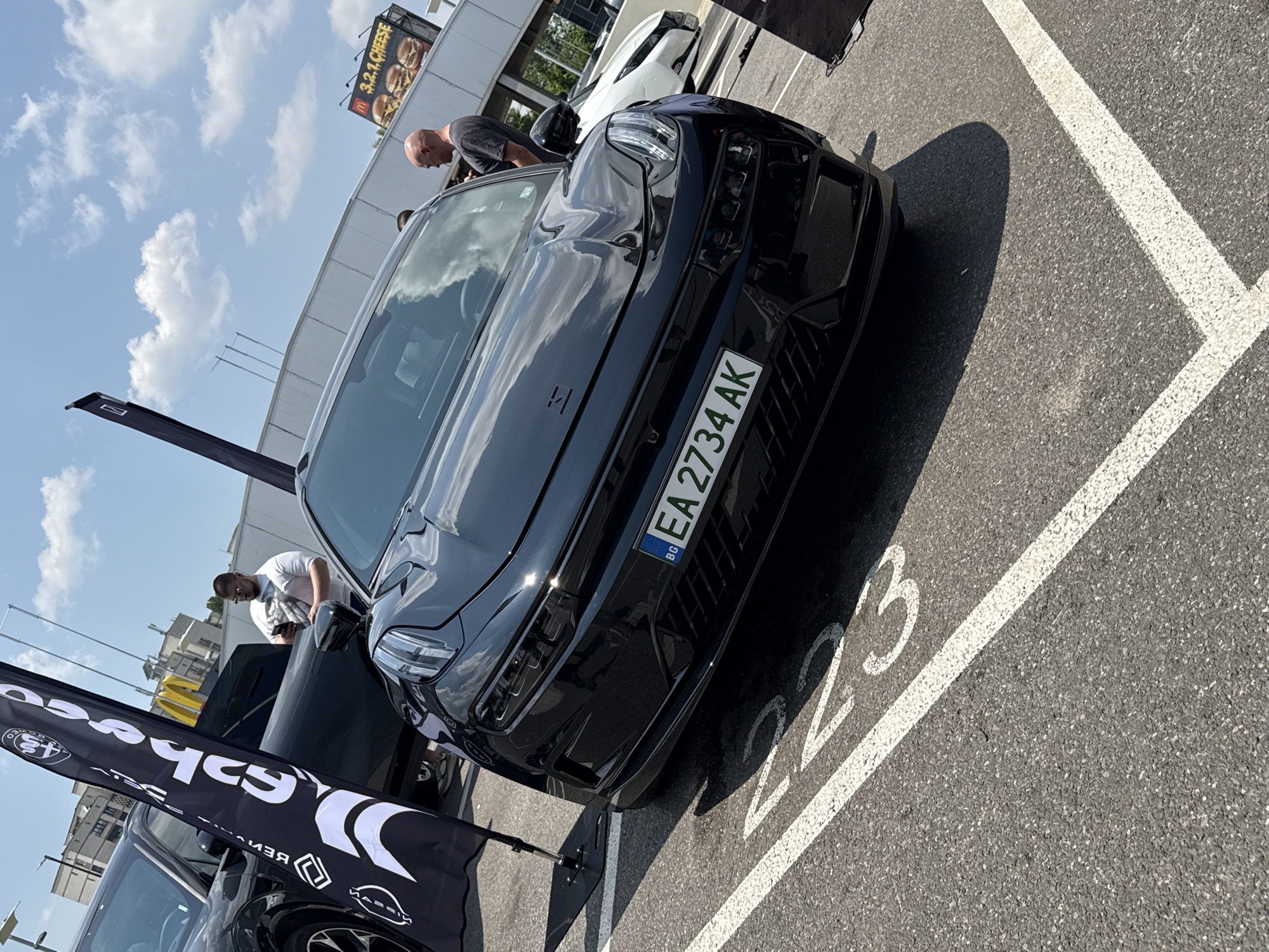 The image captures an outdoor car display or event on a sunny day in an asphalt parking lot, ostensibly located in Sofia, Bulgaria.
In the foreground, the dark grey asphalt pavement is marked with clear white lines, partially revealing the number "23" of a parking space.
The primary subject is a black Lynk & Co 03 sedan, parked at a slight angle, facing the viewer. The car displays a Bulgarian license plate: "BG EA 2734 AK." The front fascia, including the distinctive split headlights and the Lynk & Co badge on the hood, is clearly visible. Strong sunlight creates reflections on the car's glossy surface and casts a defined shadow on the asphalt to its right.
Two men are present in the midground:
1. A white male, medium build, wearing a white t-shirt and sunglasses, stands to the left of the Lynk & Co, partially obscured by a black flag. He is holding a smartphone in his right hand and appears to be observing the scene.
2. A bald white male, medium build, wearing a dark grey t-shirt, stands to the right of the Lynk & Co, looking towards the right side of the frame.
Other vehicles are partially visible: a dark-colored SUV or crossover on the far left, behind the first man and flag, and a white car (possibly a Nissan Leaf) further in the background on the right.
Two promotional flags are present: a black flag with white stylized graphics (including what appears to be reversed text "AEGIS" and arrow motifs) stands on a black base on the left, near the SUV. A small section of another black flag is visible on the far right edge of the frame.
The background features generic commercial or industrial-style buildings under a bright blue sky with scattered white cumulus clouds. A large McDonald's billboard, advertising "3.2.1 CHEESE" burgers, is prominently displayed on one of the buildings. The overall environment suggests a commercial district or shopping center parking lot.