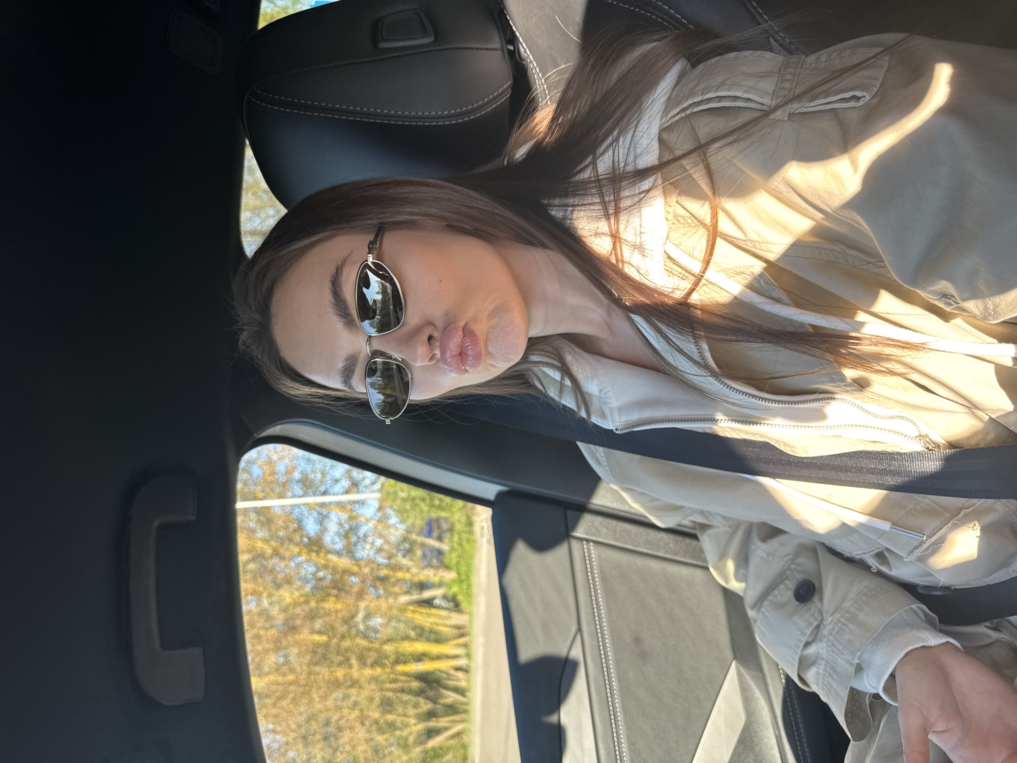 A woman wearing sunglasses and a light-colored jacket is sitting inside a car. The background visible through the car window is blurred and shows foliage and what appears to be the side of a road. The interior of the car, including the seats and a grab handle, are visible. There is no identifiable information to conclude the location is Nieporęt, Poland.