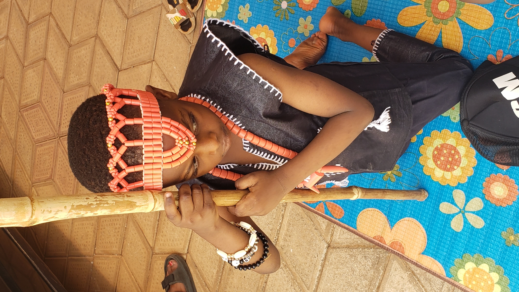 A child lies on a blue floral-patterned mat laid on a tiled surface in what appears to be Sabon Tasha, Nigeria. The child is dressed in black clothing with white trim, adorned with a beaded headpiece and necklace in shades of coral and gold. They are holding a light brown bamboo pole, and bracelets are visible on their wrist. A black baseball cap rests near the edge of the mat. Footwear, including sandals, are also visible near the mat.
