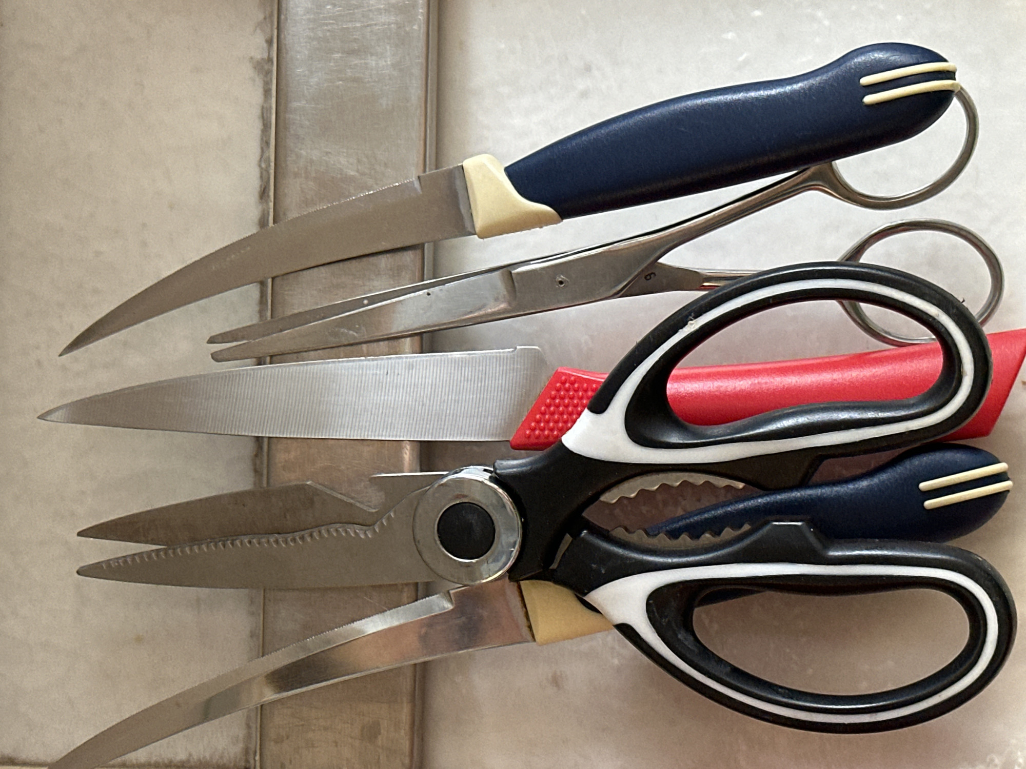 The image shows a close-up, eye-level view of several kitchen shears and knives hanging on a magnetic strip against a light-colored, textured wall. The objects are arranged from left to right.
On the far left, there is a silver carving knife with a long, narrow blade and a yellow handle. Next to it are a pair of black and white handled kitchen shears, with one shear blade visible and a serrated edge. Attached to these shears, and slightly overlapping, is a red plastic piece, possibly a safety cover or part of the handle. To the right of that is another pair of kitchen shears, these with silver blades and silver handles, with the number "9" faintly visible on one of the handles. To the far right is a knife with a silver blade and a dark blue handle with two yellow accents near the bolster.
The background appears to be a tiled or stone wall with some subtle variations in color and texture, suggesting a kitchen environment. The lighting is bright and even, with no strong shadows. The image is a still life, focusing on the tools.
There are no people, animals, or specific identifiable landmarks or city features visible in the image. Therefore, it is impossible to determine the location in the city of Nevinnomyssk, Russia, or any other specific geographic location.