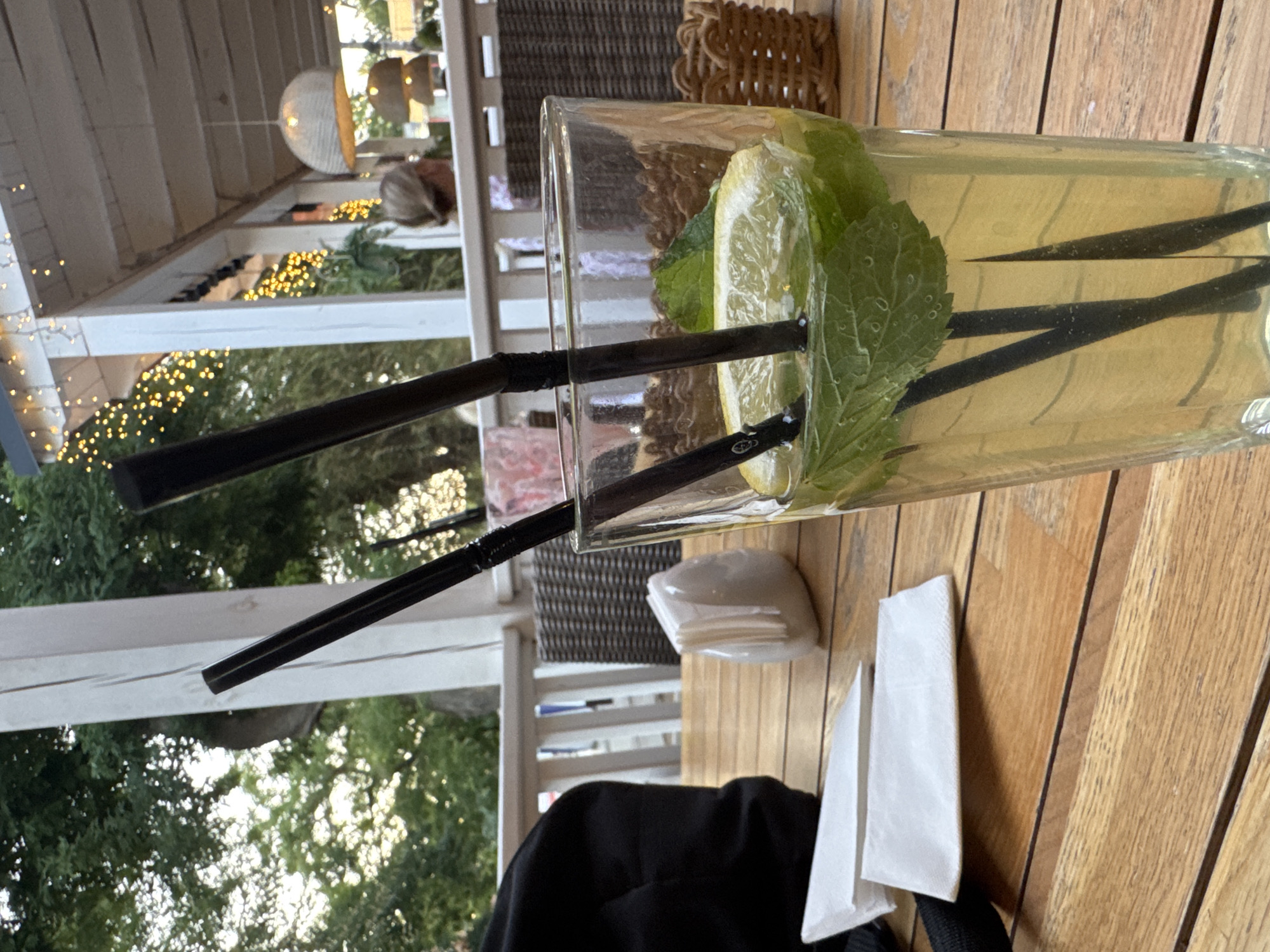This is a medium shot, eye-level, indoor/outdoor photograph of a drink on a wooden table. The drink, likely lemonade or iced tea, is in a clear glass and contains ice, a slice of lemon, and mint leaves. Two dark straws are in the glass. The table is made of light-colored wood planks. To the left of the drink are two white paper napkins, folded and stacked. Further left is the black fabric of what appears to be a bag. Behind the table, a white railing separates the area from a walkway or outdoor seating space. In the background, foliage from trees is visible, along with the exterior of a building with white columns and a light-colored roof. Decorative yellow lights are strung along the roofline. A person's hair is visible over the railing in the background, suggesting other patrons are present. The scene appears to be at a cafe or restaurant, possibly in Yevpatoriya, Ukraine, given the environmental context.
