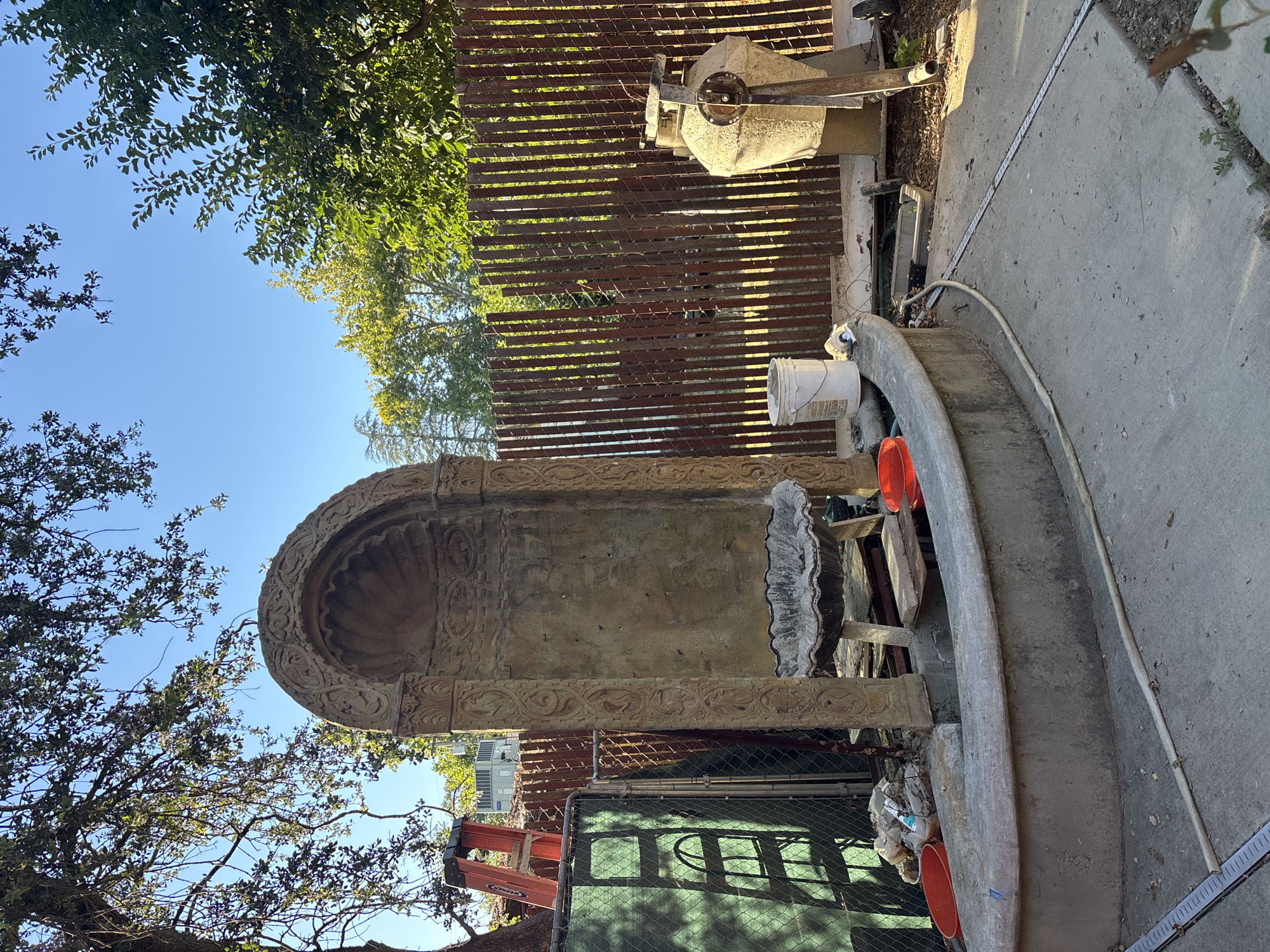 An outdoor scene in Fair Oaks, California, depicts a large, weathered concrete fountain in a state of disrepair. The fountain's central element is an arched structure with intricate carvings, resembling a niche or grotto. Below this, a lower basin is visible, also made of concrete and showing signs of age. Two bright orange plastic buckets are placed near the fountain's edge, one partially filled with debris. A white bucket with what appears to be construction material sits on the ground beside a rustic wooden fence. To the left of the fountain, a green construction screen is draped over a chain-link fence, with an orange ladder leaning against it. Beyond the fence, trees with lush green foliage and a clear blue sky are visible. A large, cream-colored urn-like structure with a metal mechanism attached stands to the right of the fence. The ground is paved with concrete, with some debris and fallen leaves scattered around. The overall impression is one of a construction or renovation project underway in a backyard setting.