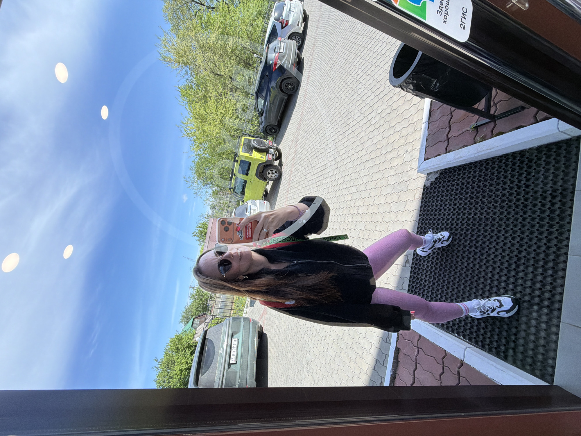 A woman is standing in front of a window, taking a selfie with a phone with a photo on the screen. She is wearing sunglasses, a black jacket, and pink leggings with white sneakers. Outside the window, there are several parked cars, including a green Jeep and a silver Hyundai. The pavement is visible, and trees are in the background. There is a sign in Russian that says "Здесь хорошо 2ГИС" which could indicate that the location is in Russia. Given the cars and the sign in Russian, it is possibly somewhere in Russia, and there is a higher probability of the city Birobidzhan.