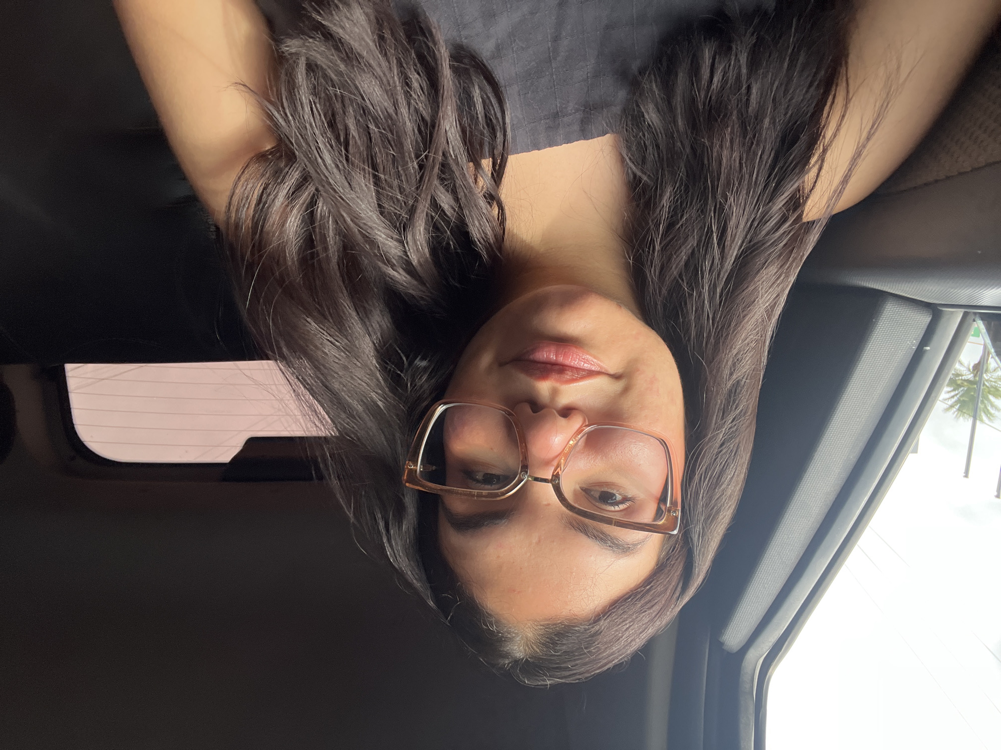 A young woman with dark, wavy hair and glasses is seated in the back of a vehicle, likely a car or van. The interior of the vehicle appears dark, with black upholstery visible. Sunlight streams in from the left side, illuminating the woman's face and hair. Through the window, a blurred outdoor scene is visible, suggesting the vehicle is in motion. The woman is wearing a dark, sleeveless top. Her facial features are clear, and she appears to be looking directly at the camera. The image does not provide any specific visual cues to pinpoint the location within Porlamar, Venezuela.
