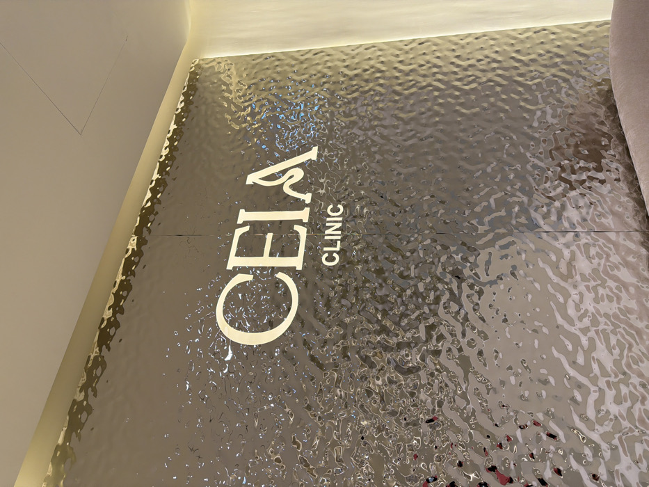A tilted view of a textured, possibly metallic, surface. The words "CELA CLINIC" are prominently displayed in a vertically stacked arrangement on the left side, in a light color, possibly gold or cream. The texture of the background resembles a shimmering or rippled pattern, creating an interesting visual effect. The lighting suggests it is indoors.