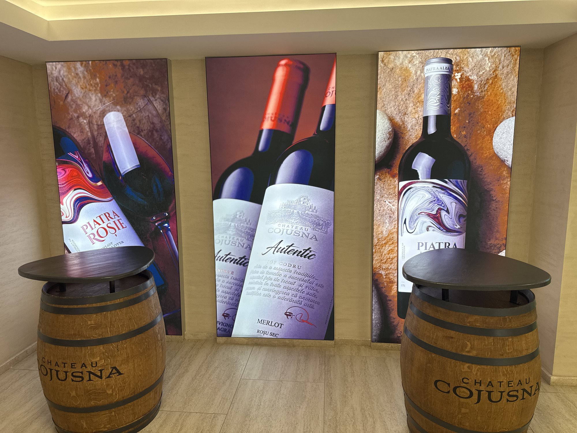 The image shows a well-lit interior, possibly a tasting room or retail space, featuring three large vertical posters depicting wine bottles and labels. In the foreground, two wooden wine barrels are positioned as tables, each topped with a dark, circular surface. The posters display a consistent theme with prominent branding for "Chateau Cojușna" and various wine names such as "Piatra Rosie," "Autentic," and "Piatra." The lighting highlights the glossy texture of the wine bottles and the stone-like backgrounds in the posters. The overall ambiance suggests a sophisticated presentation of wine products, likely associated with the Chateau Cojușna winery in Cojușna, Moldova.