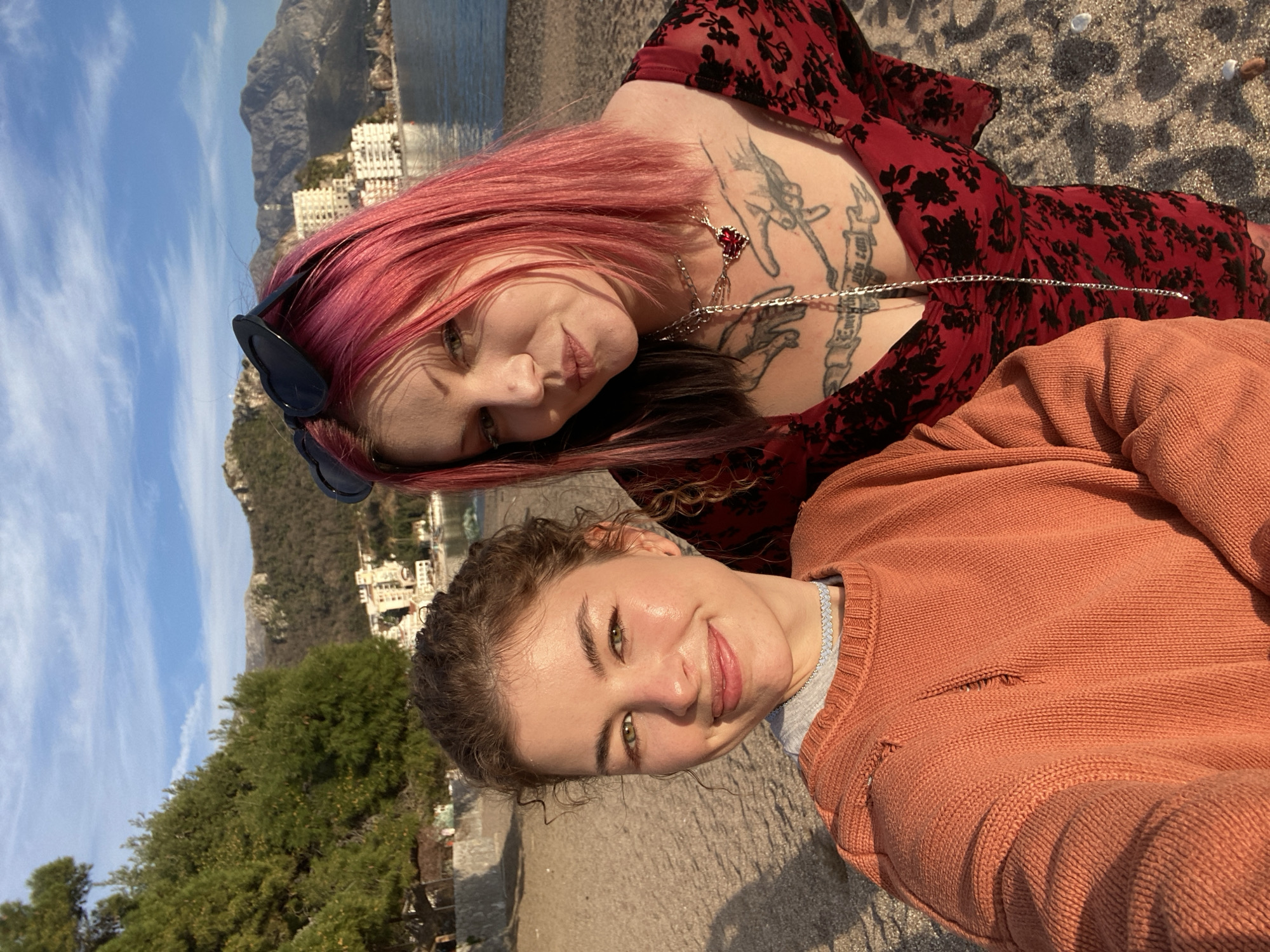 Two females are depicted in a selfie-style photograph on a sandy beach in Rafailovići, Montenegro. The individual on the left, with light brown curly hair, green eyes, and an orange-rust distressed knit sweater over a grey undershirt and a light choker, smiles at the camera. The individual on the right, with pink hair, dark roots, and heart-shaped sunglasses pushed onto her head, wears a red low-cut dress with black floral patterns, revealing chest tattoos of two reaching hands and text, adorned with multiple necklaces including a silver chain and a red heart pendant; she looks directly at the camera. The background shows a calm blue sea, a coastal settlement featuring multi-story white buildings, and a range of mountains with varied vegetation under a partly cloudy blue sky. Lush green trees are visible on the left, bordering a low concrete wall along the beach. The beach sand contains scattered small pebbles. Natural sunlight illuminates the scene, casting visible shadows on the sand.
