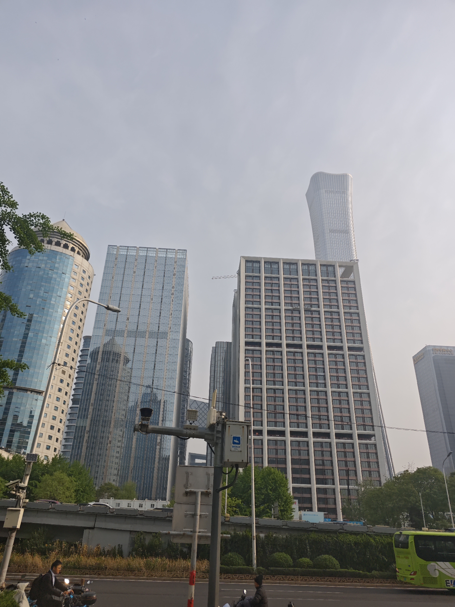 The image captures a street scene in Chaoyang District, Beijing, China. In the background are multiple skyscrapers with a mix of glass and solid facades, including the prominent CCTV Headquarters building. A surveillance camera system is prominently featured. In the foreground, two people are driving on motorbikes on the street. A green bus is on the right side of the image. The sky is overcast.