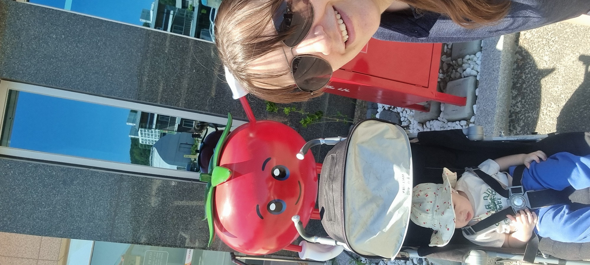In Dunnae-myeon, South Korea, a woman takes a selfie outdoors with a child in a stroller and a tomato statue. The woman wears sunglasses and a gray top. The child lies in the stroller, strapped in, and wears a hat. The red tomato statue has a smiling face and green leafy stems. The scene includes a building with reflective windows, a red metal structure, white gravel, and a stroller with a gray sunshade.