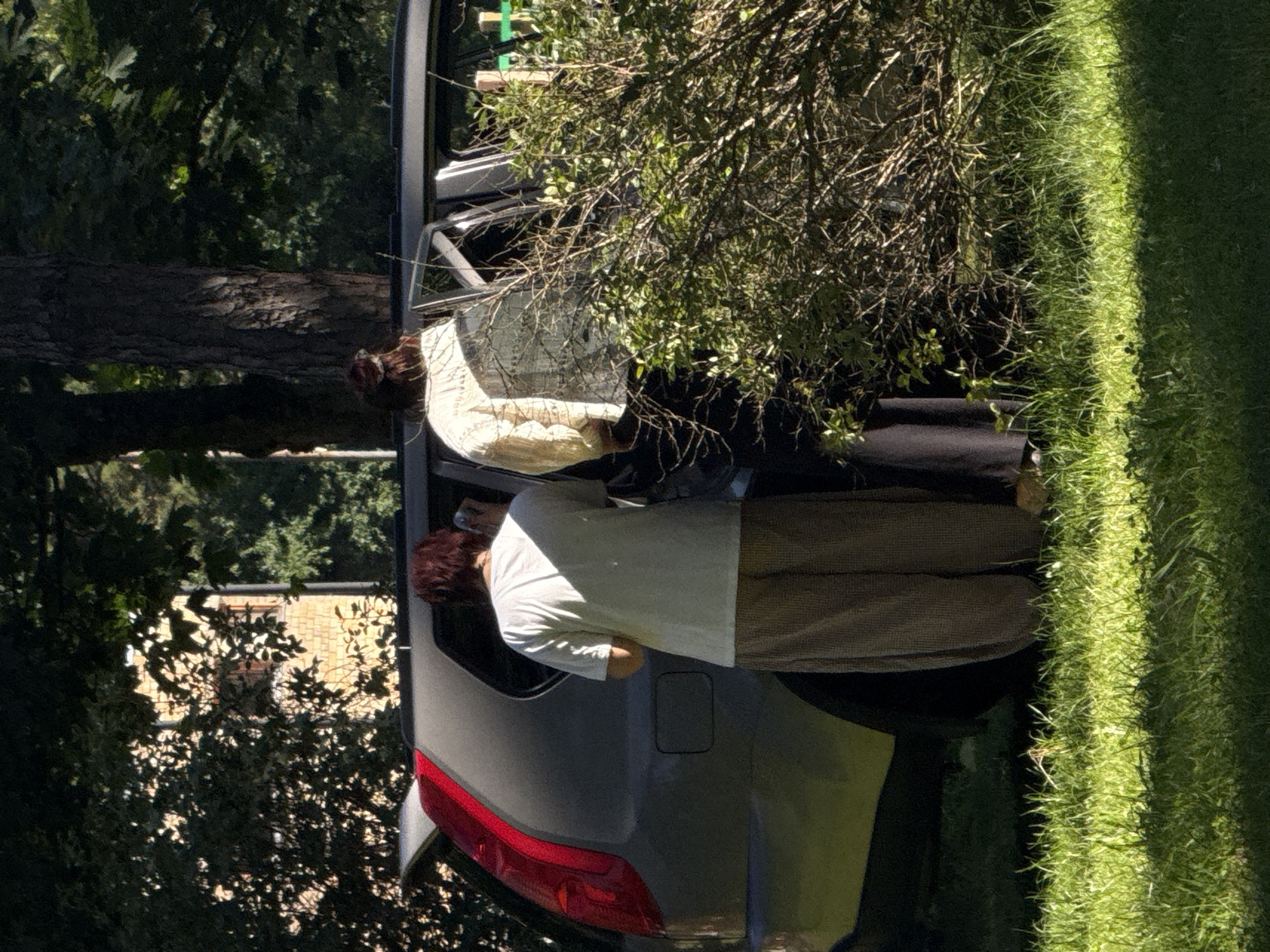 Two individuals, one wearing a white t-shirt and brown checkered pants, and the other wearing a cream-colored patterned blouse and dark pants, stand beside an open car door. The car, a silver SUV with its trunk open, is parked on a grassy area with dappled sunlight. Behind the car, trees and foliage create a natural, outdoor setting. A building with a shingled facade and visible windows is partially obscured by trees in the background. The scene appears to be in an outdoor, residential or park-like environment, possibly in the city of Ventspils, Latvia, given the style of the building and the overall natural landscape.
