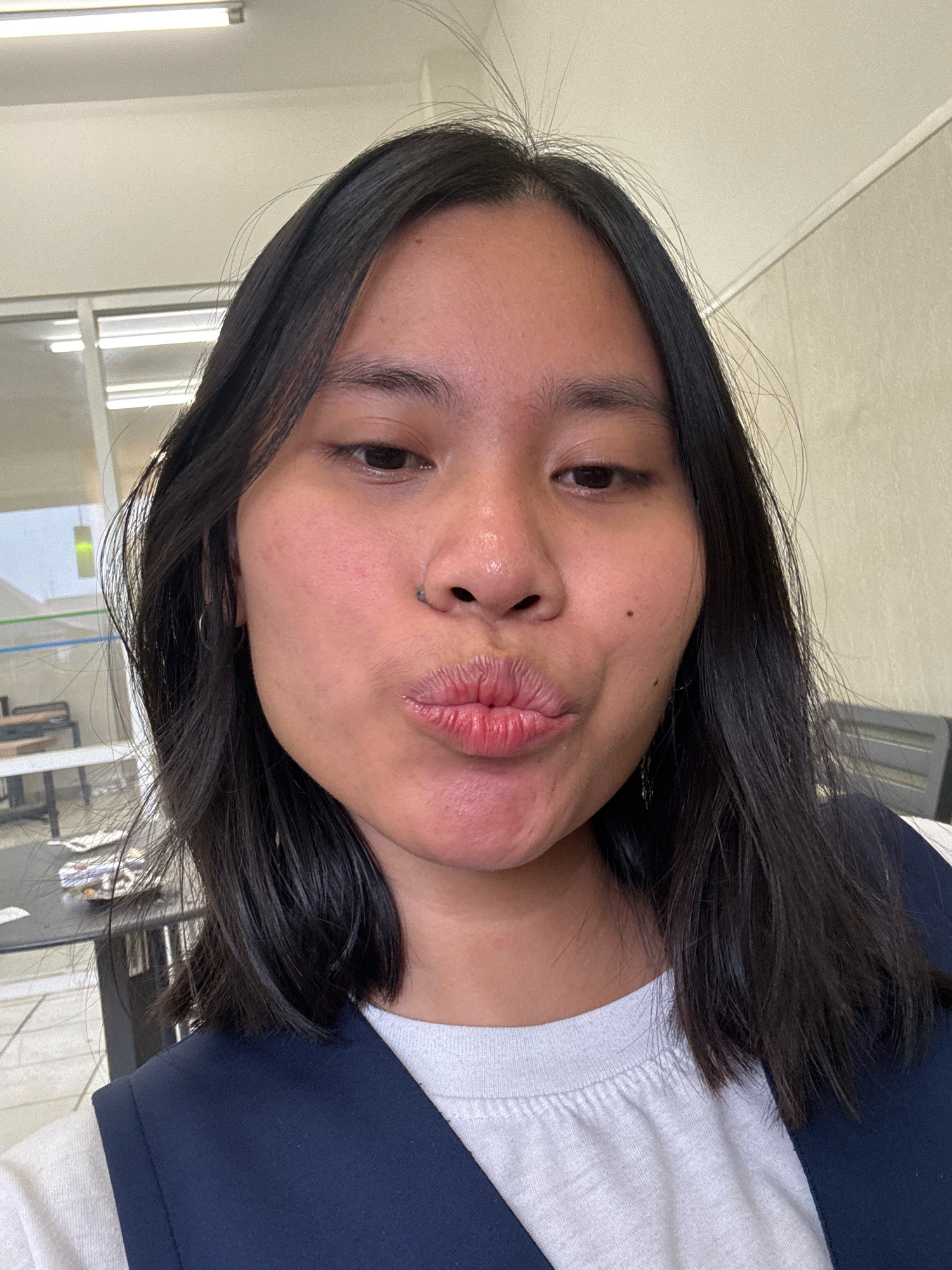 The image is a close-up selfie of a young East Asian woman with dark hair, puckering her lips for a kiss. She is wearing a white t-shirt and a dark blue vest. She has a nose ring. The background appears to be an indoor setting with fluorescent lighting on the ceiling, large windows with a view of what looks like an office or a school, and some furniture visible. The lighting suggests an indoor environment, possibly an office or educational institution. The overall impression is a casual selfie taken indoors.