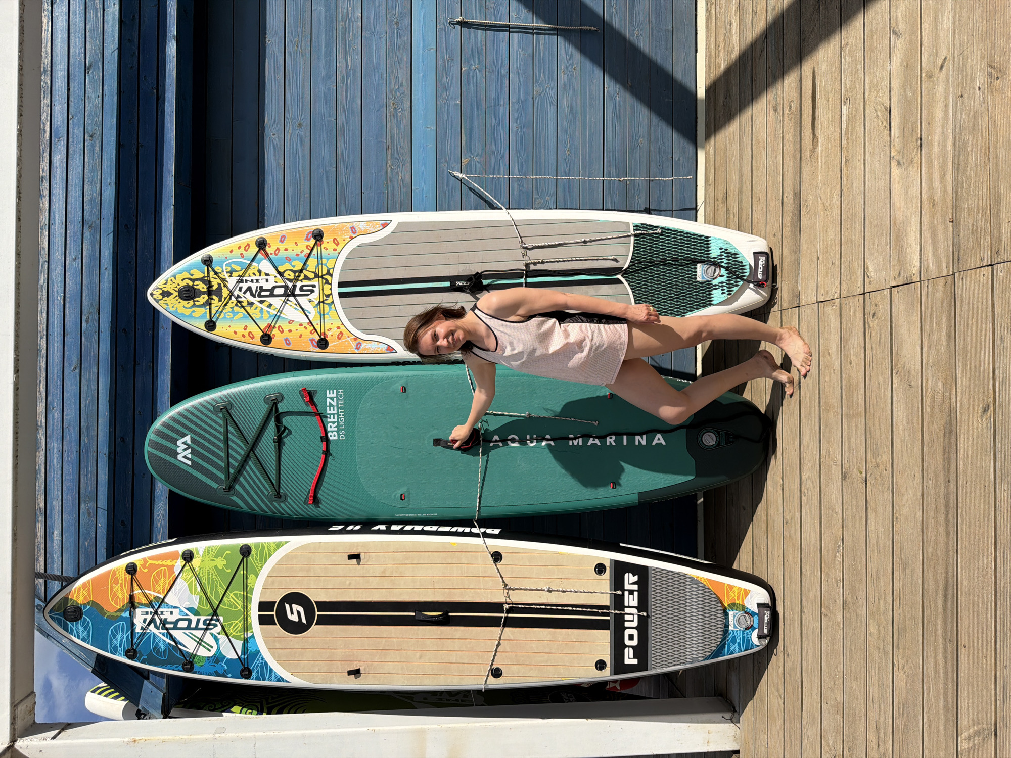 The image shows a woman lying on three stacked paddleboards on a wooden dock or platform. The boards are of different brands and designs. The woman is wearing a light-colored tank top and has bare feet. The dock is bordered by a blue wall or structure. The location is Chaikovsky, Russia.