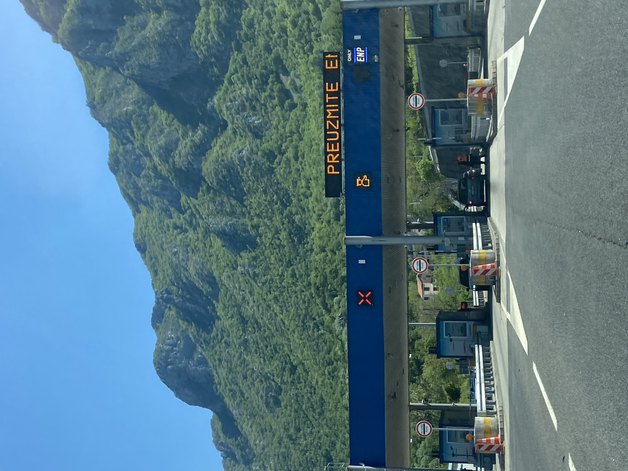 The image depicts a toll booth on a road, likely a highway, in Gluhi Do, Montenegro. The mountains are to the left, the road to the right. The booths have traffic lights, some with red "X" signs, and signage above indicating payment options. "PREUZMITE E1" is displayed on the lighted signage, as well as "ONLY ENP". There is one car waiting at the booths. Signs saying “hit a horn please” are also visible. The sky is blue and the mountains are green.