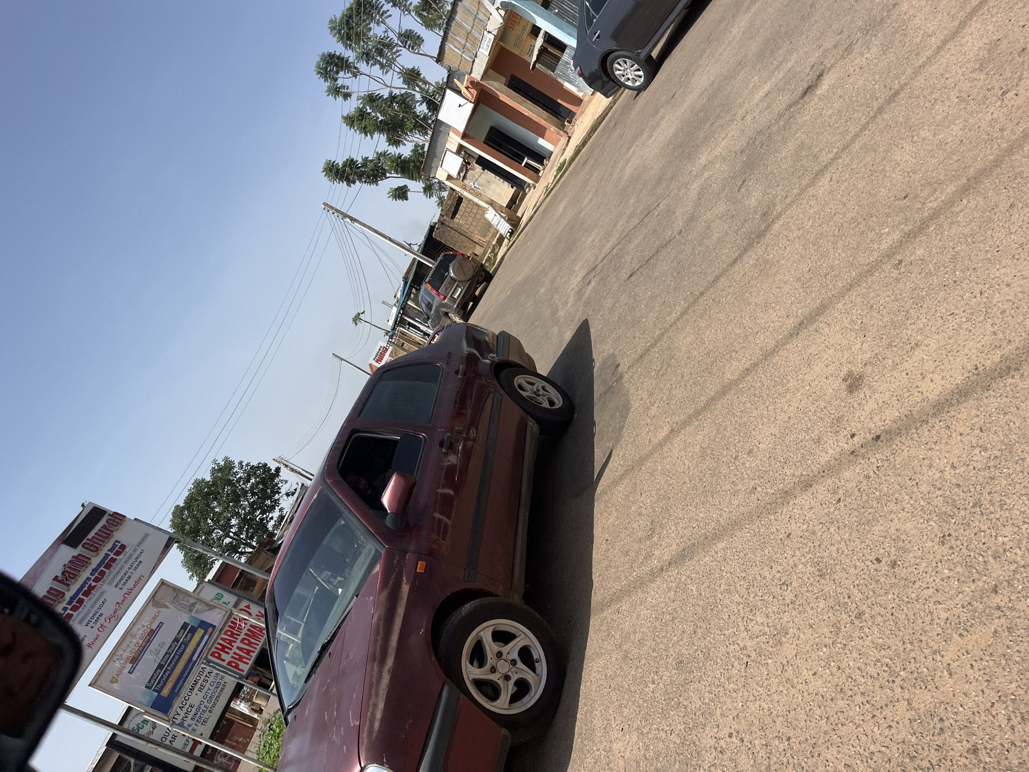The image depicts a street scene in Gura Top, Nigeria. In the foreground, a maroon Volkswagen Golf hatchback is parked on the right side of the road. Further down the street, an SUV and a dark gray sedan are also visible. Several small shops with colorful facades line the right side of the road. A sign for "Living Faith Church" is prominently displayed. The sky is clear and blue. The road surface is asphalt.