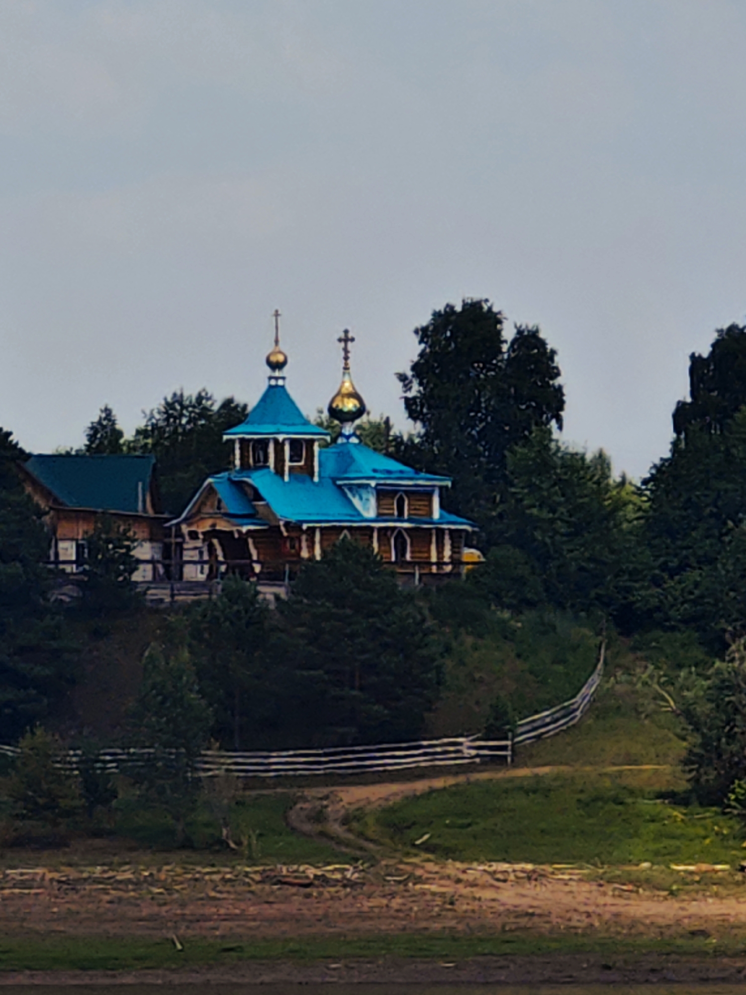 A wooden church with bright blue roofs and golden domes sits on a hill, surrounded by dense green trees. A white wooden fence lines a winding dirt path leading up the hill. The sky is overcast and gray. The image appears to be taken in Erkalovo, Russia.