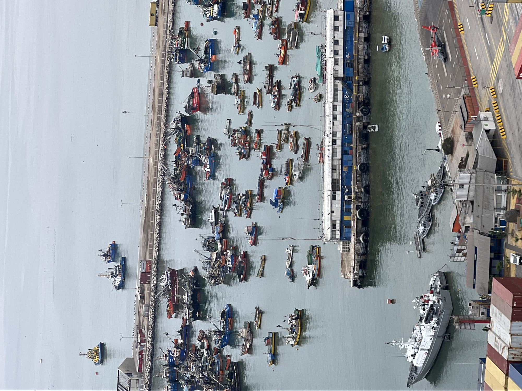 This high-angle image depicts a bustling port and harbor scene in the city of Arica, Chile, under an overcast sky. The foreground reveals a section of port infrastructure, including a gray patrol boat identified as "ARICA" on its stern, docked near a concrete quay. A smaller rigid-hull inflatable boat (RHIB) is stowed on the patrol boat's aft deck. Adjacent to the quay, a helipad is visible with a red and black helicopter (likely a Bell 412 or similar) parked on it. Various shipping containers (red, yellow, blue) and low-rise port buildings line the waterfront, with a white pickup truck on an asphalt road featuring yellow and black striped barriers.
The midground is dominated by an inner harbor densely packed with numerous small to medium-sized fishing vessels, predominantly trawlers or purse seiners, displaying various colors including blue, red, yellow, and white. These boats are rafted together or individually moored, many equipped with derricks, outriggers, and nets. A prominent blue and white floating pier or administrative building is moored in the central midground. Several smaller boats, buoys, and scattered birds are also present in this area.
Separating the inner harbor from the open sea is a long, concrete breakwater or pier. Beyond this structure, in the background, a vast expanse of calm, gray water extends under the overcast sky. A yellow and blue tugboat is visible in the upper left, with two blue fishing vessels further out in the upper right. A solitary dark buoy is discernable in the distant water. The overall scene indicates a busy commercial and fishing port operation.