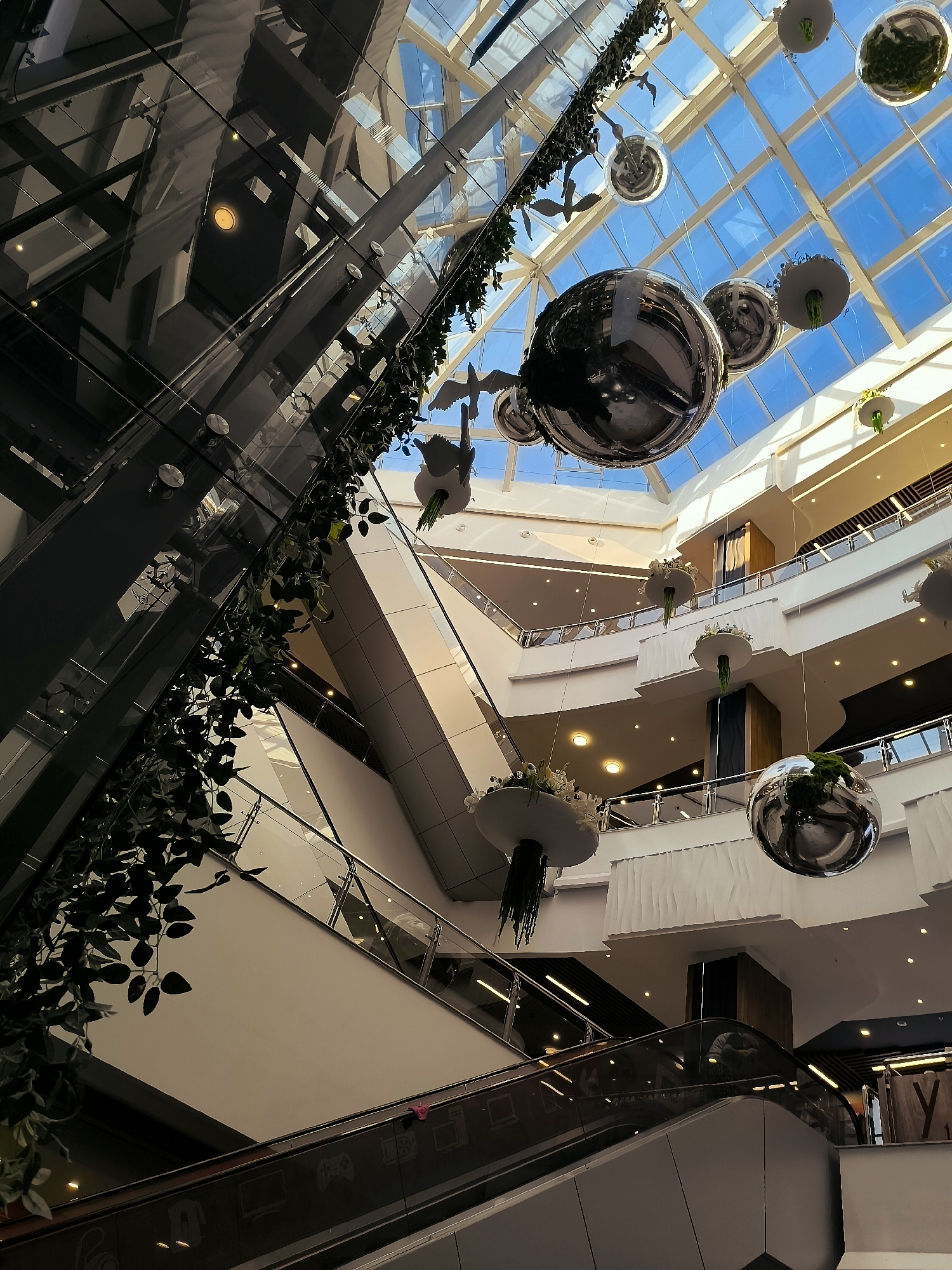 This image depicts the interior of a multi-level shopping mall with a glass ceiling. The architecture features sweeping escalators, balconies with glass railings, and a central atrium. Decorative elements include large, reflective spherical ornaments and hanging artificial greenery. The overall aesthetic is modern and spacious, with natural light filtering through the ceiling. The scene is devoid of any discernible people or specific brand signage, presenting a generic mall environment. There is no specific visual information in the image to indicate it is located in the city of Qarağandı city administration, Kazakhstan.