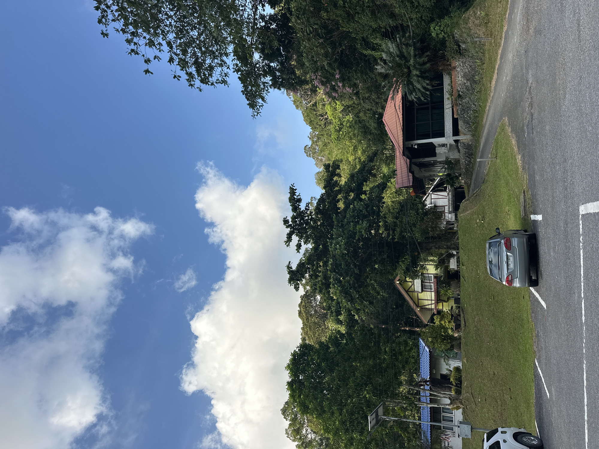 A scenic view captures a residential area in Tambon Wang Prachan, Thailand, nestled at the base of a lush, tree-covered hillside under a bright blue sky dotted with fluffy white clouds. In the foreground, a paved parking lot with white markings indicates a designated parking area. A silver sedan is parked in one of the spots, facing away from the camera, while the rear of a white SUV is visible on the left.
A gently sloping grassy embankment leads up to two houses. The house on the left features a blue roof and white walls, with a solar panel on a pole situated in front of it. Adjacent to this, a yellow house with a brown-tiled roof displays brown trim around its windows. Further up the slope and to the right, a larger house with a prominent red-tiled roof is visible, supported by a stone foundation. A paved road curves away from the parking lot, leading towards this larger house. The environment is characterized by abundant greenery, with dense trees forming a natural backdrop to the houses and a feeling of tranquility.