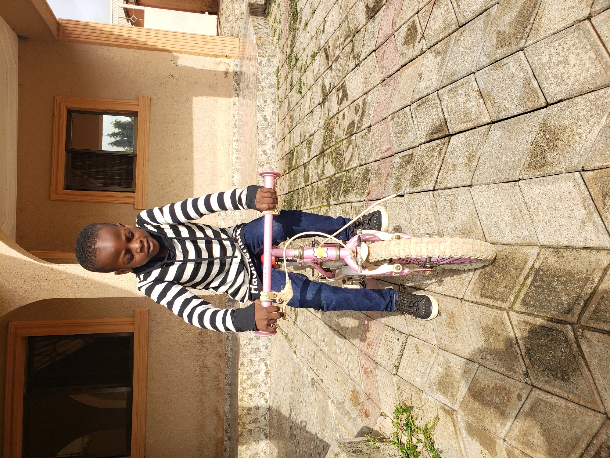 A young Black boy is seated on a pink bicycle on a paved outdoor area in front of a building. He is wearing a black and white striped hoodie and dark blue jeans, with black sneakers. The bicycle has training wheels and a pink frame. The background features a light brown building with two windows and arched doorways. The ground is paved with interlocking stone tiles, some of which appear wet. The scene is lit by daylight, casting shadows on the ground. The location is identified as Sabon Tasha, Nigeria.