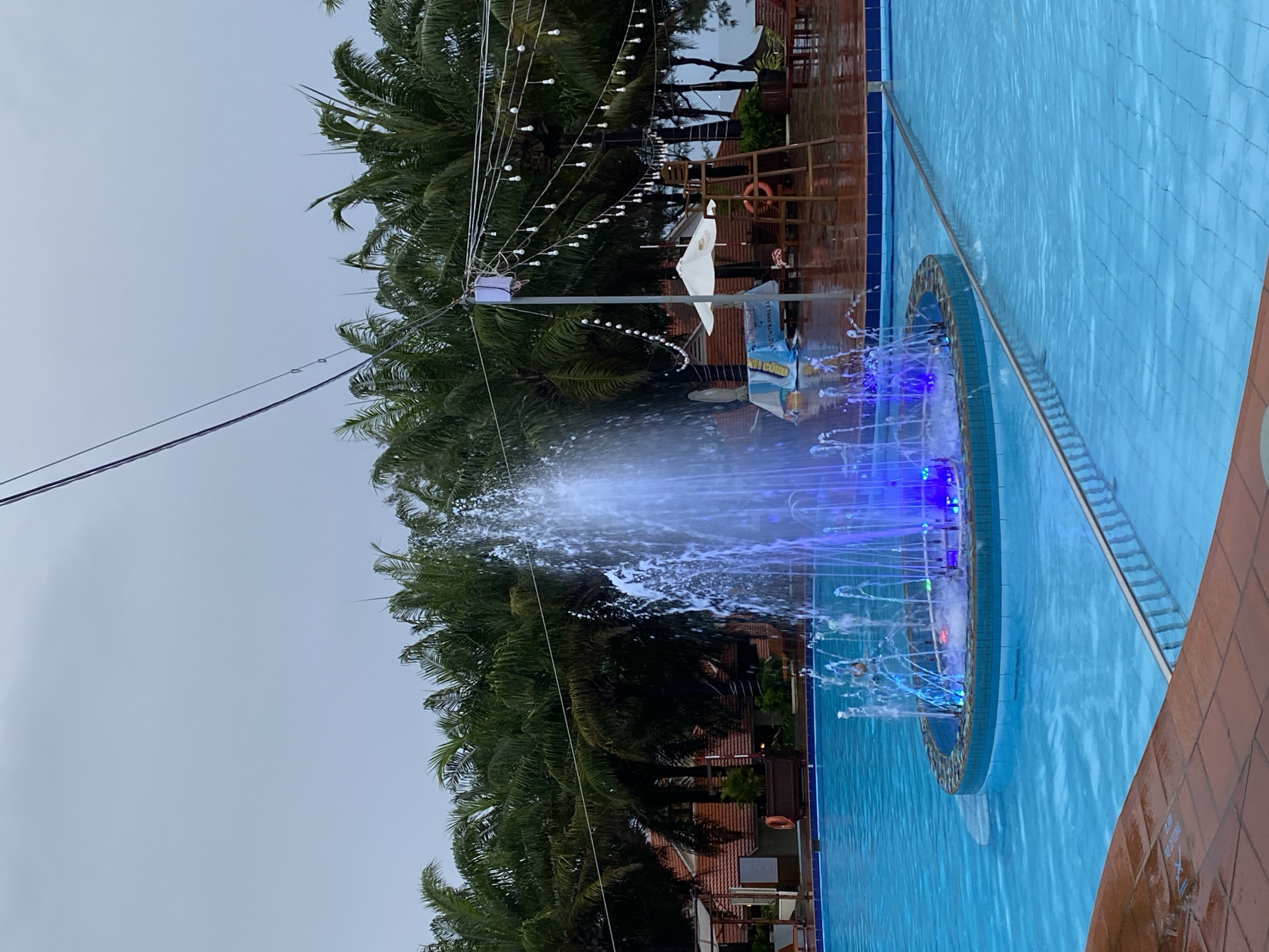 The image shows a fountain in a swimming pool in Phu Thuy Ward, Vietnam. The fountain is circular, with blue lights illuminating the water jets. The pool is tiled in blue and surrounded by brickwork. Palm trees and building are in the background. String lights are hung above the scene. The sky is overcast.