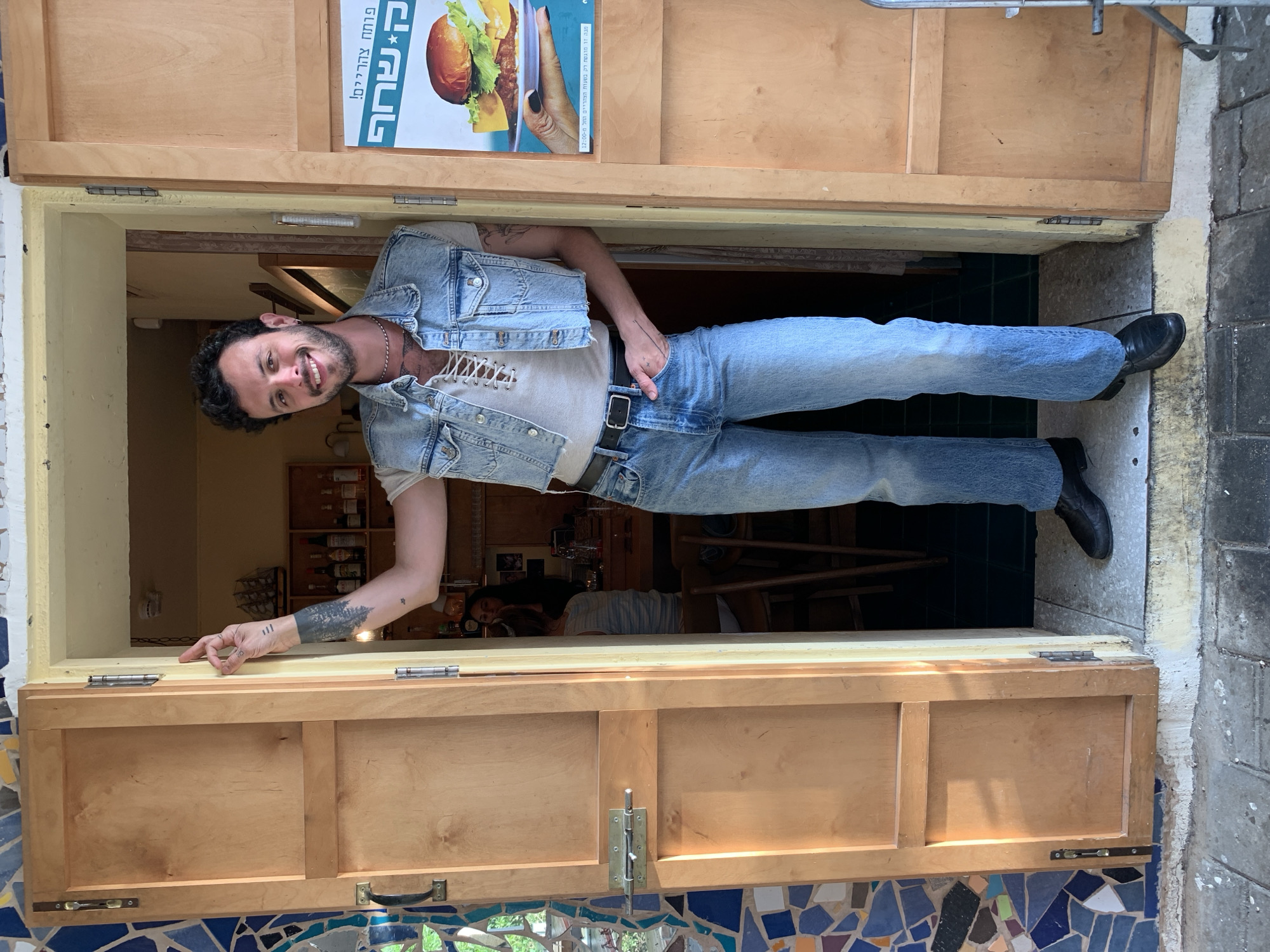 A man dressed in denim clothing stands in a doorway in Tel-Aviv, Israel. The man wears a denim vest over a light-colored shirt and denim jeans, cinched with a black belt, and black shoes. He has dark hair and a beard. A tattoo is visible on his left arm. The doorway's construction is of light-colored wood, accented with blue and multi-colored mosaic tiles around its base. A sign featuring a burger image and Hebrew text is posted at the top of the doorway. The Hebrew text says, "Opens at noon! Kashtan" (קשחף) - probably the name of the restaurant. Other people and objects can be seen within the indoor space behind the doorway, including shelves stocked with bottles.