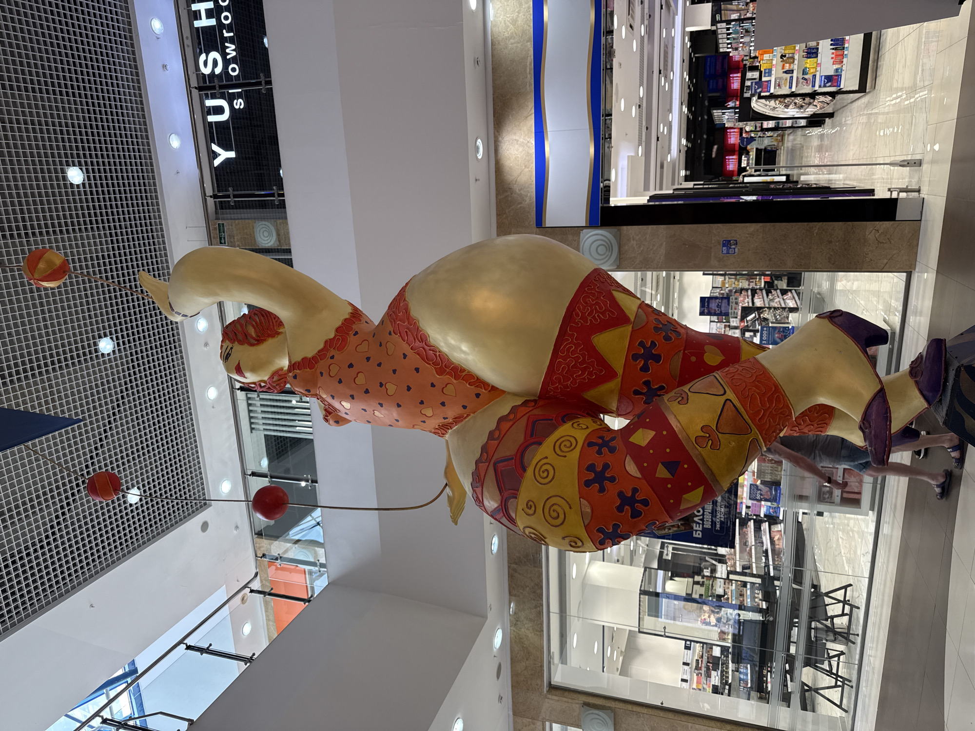 The image shows a large, colorful statue of a circus performer in what appears to be a shopping mall in Nizhny Novgorod, Russia. The statue is of a female figure with golden skin, wearing a red and orange patterned outfit. She is holding two red and orange balls attached to strings. The environment is a modern shopping mall with multiple levels, glass railings, and brightly lit retail spaces. In the background, there is a store with the word "YUSH Showroom" visible. There are people visible in the background, walking around the mall. The statue seems to be a decorative feature of the shopping center.