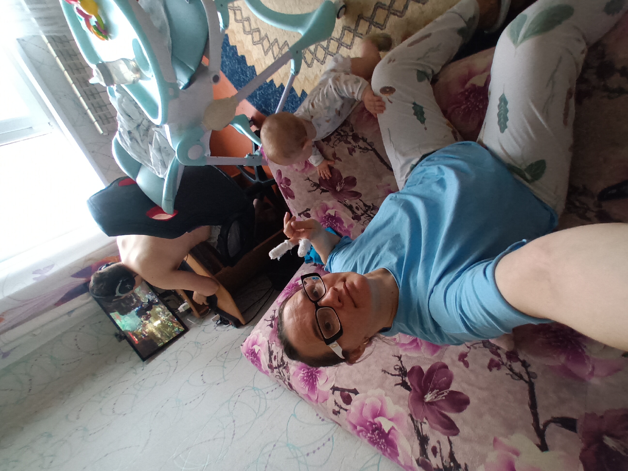 The image depicts a woman lying on a bed with a floral patterned pillow in a room with a patterned wall and a rug on the floor. She is wearing glasses and a blue shirt, and holding a white object in her right hand. A baby lies on the bed nearby, close to a baby activity center. Another person is visible in the background near a television. The location is Ольховка, Russia.