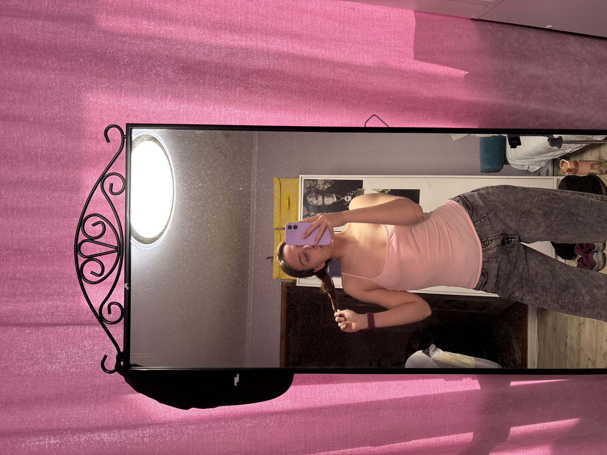 A young woman is taking a mirror selfie. She is wearing a pink tank top and grey jeans. The mirror is rectangular with a decorative wrought iron frame. The background is a pink curtain or backdrop. The location is not definitively discernible as Прикамский, Russia from the available visual information.