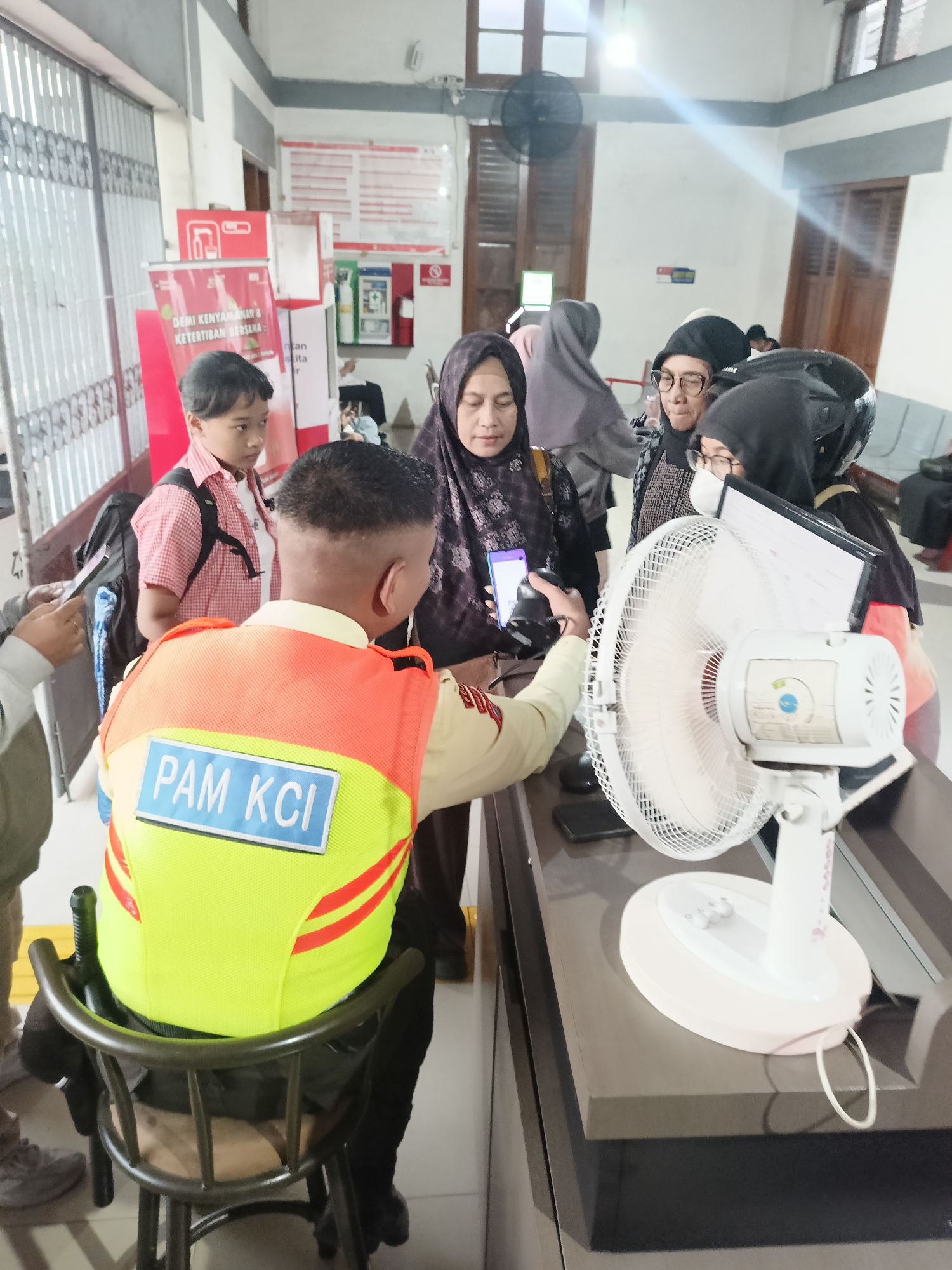 In Gedangan, Indonesia, a security guard wearing a bright yellow and orange vest with "PAM KCI" written on the back is seated on a stool. He is overseeing a line of people, including a young boy in a red and white checkered shirt with a backpack, and several women wearing hijabs. One woman is holding a smartphone, possibly scanning a ticket or boarding pass. A white electric fan is positioned on a counter in the foreground. The environment appears to be an indoor waiting area, possibly a train station, with posters and ticket machines visible in the background. The lighting suggests an indoor setting with overhead lights contributing to the illumination.