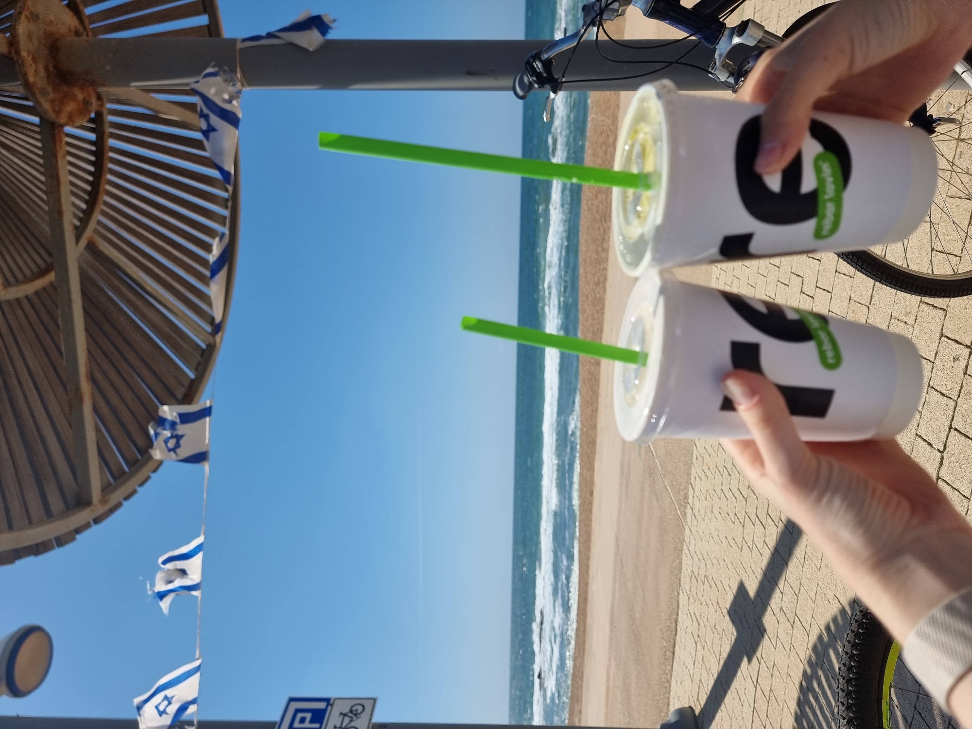 The image depicts a sunny beach scene, likely in Tel Aviv, Israel, based on the presence of Israeli flags in the background. The sky is clear and blue, and the ocean stretches out to the horizon, where the sky meets the sea. Waves are visible crashing on the sandy beach.
In the foreground, a person is holding two white cups with green straws. The cups have a logo on them. A bicycle is also visible, parked nearby. The ground is paved.
The scene is bright and suggests a warm, sunny day at the beach, with a person enjoying refreshments while on a bike ride.