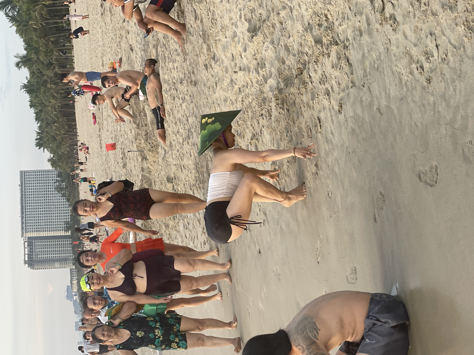 This image depicts a crowded beach scene in Đà Nẵng, Vietnam. Several groups of people are visible, engaging in various activities. In the foreground, a person wearing a conical hat, possibly a non la, and a white top with black shorts, is bent over with their hands touching the sand. To the left, a group of women in swimwear are walking along the beach, some carrying bags. In the middle ground, several men are sitting or lying on the sand, some appearing to be relaxed or socializing. In the background, more people are scattered across the beach, and a row of palm trees lines the edge of the sand, beyond which are buildings, suggesting an urban coastal setting. The sand is a prominent feature of the environment, with footprints visible in several areas. The sky is overcast, casting a soft light over the scene.