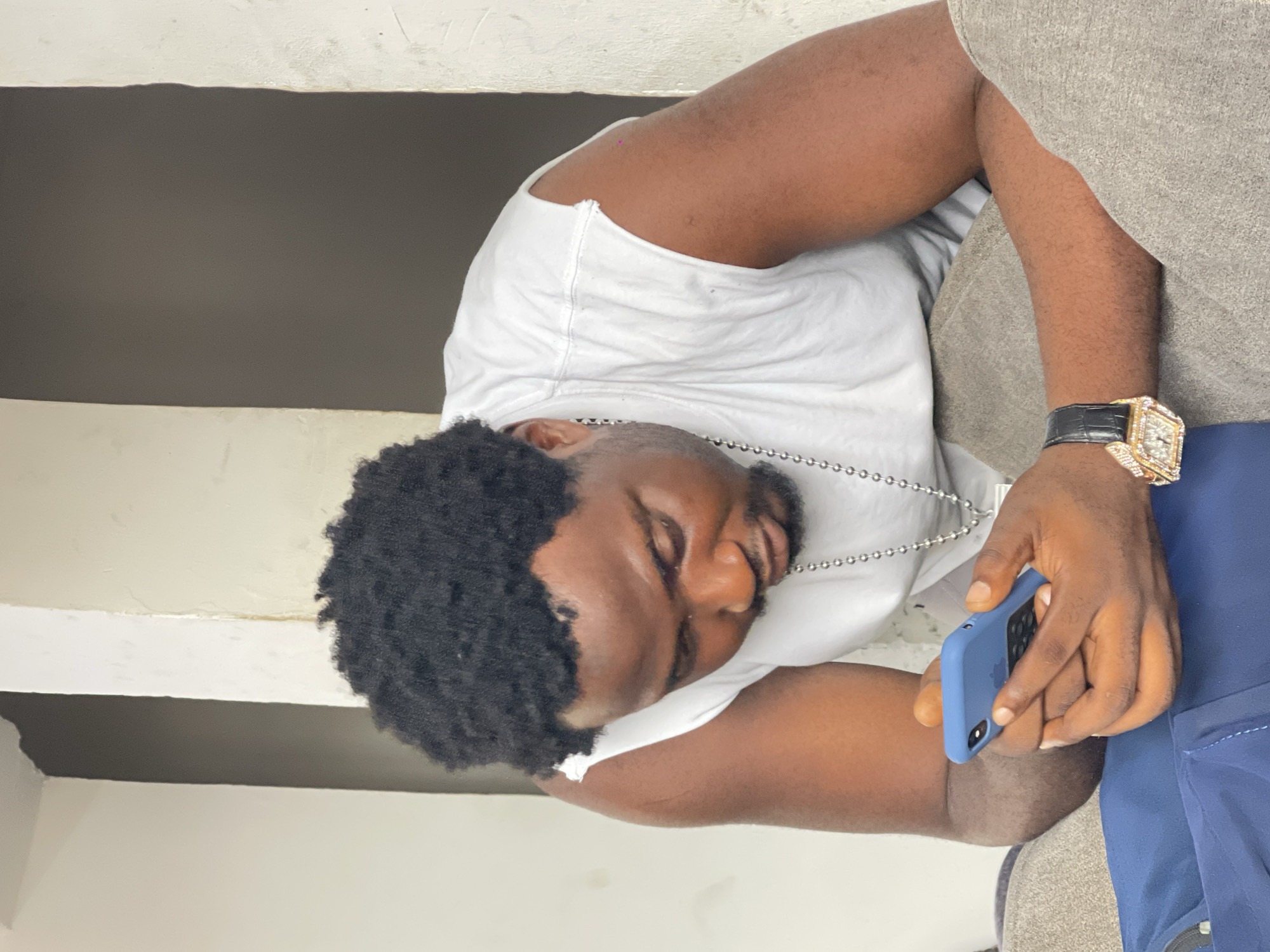 A man with dark, curly hair is seated on a couch, looking down at his blue smartphone. He is wearing a white sleeveless shirt and a silver chain. On his left wrist, he wears a gold watch with a black band, adorned with numerous diamonds. The background consists of a light-colored wall with a darker vertical section. The man's pose suggests he is engrossed in his phone. The setting is likely an indoor residential space in Abuja, Nigeria.