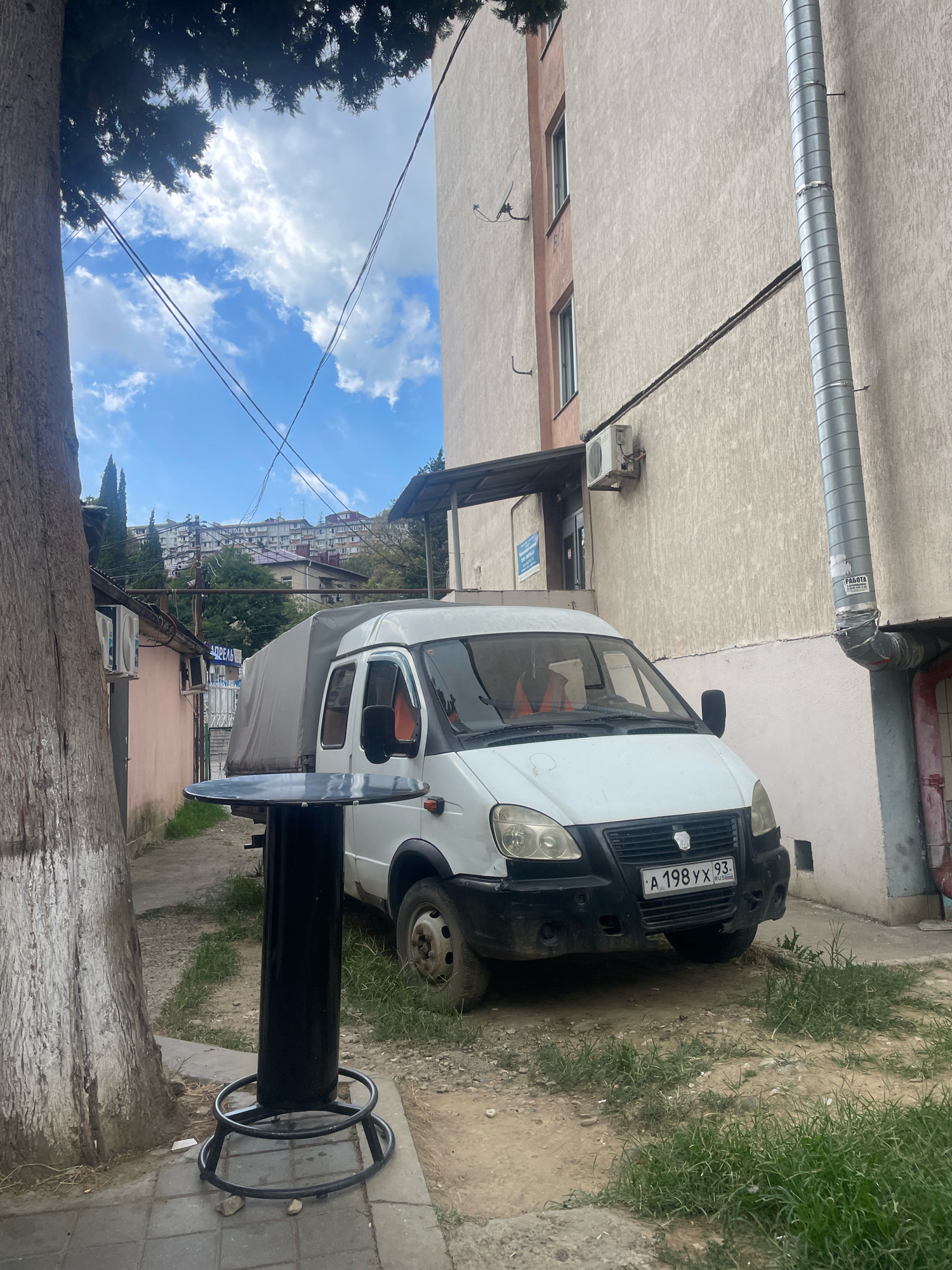A white van with a gray tarp covering its cargo area is parked on a dirt path next to a light-colored apartment building. The van has a black grille and bumper, and a license plate reading "A 198 YX 93 RUS". A person wearing an orange safety vest is visible in the driver's seat. In the foreground, a tall black cylindrical table with a round top and a circular base sits on a paved walkway. To the left of the table is the trunk of a large tree with rough bark. The background shows more apartment buildings, trees, and power lines under a partly cloudy sky. The scene appears to be in a residential area of Sochi, Russia, possibly a courtyard or alley.