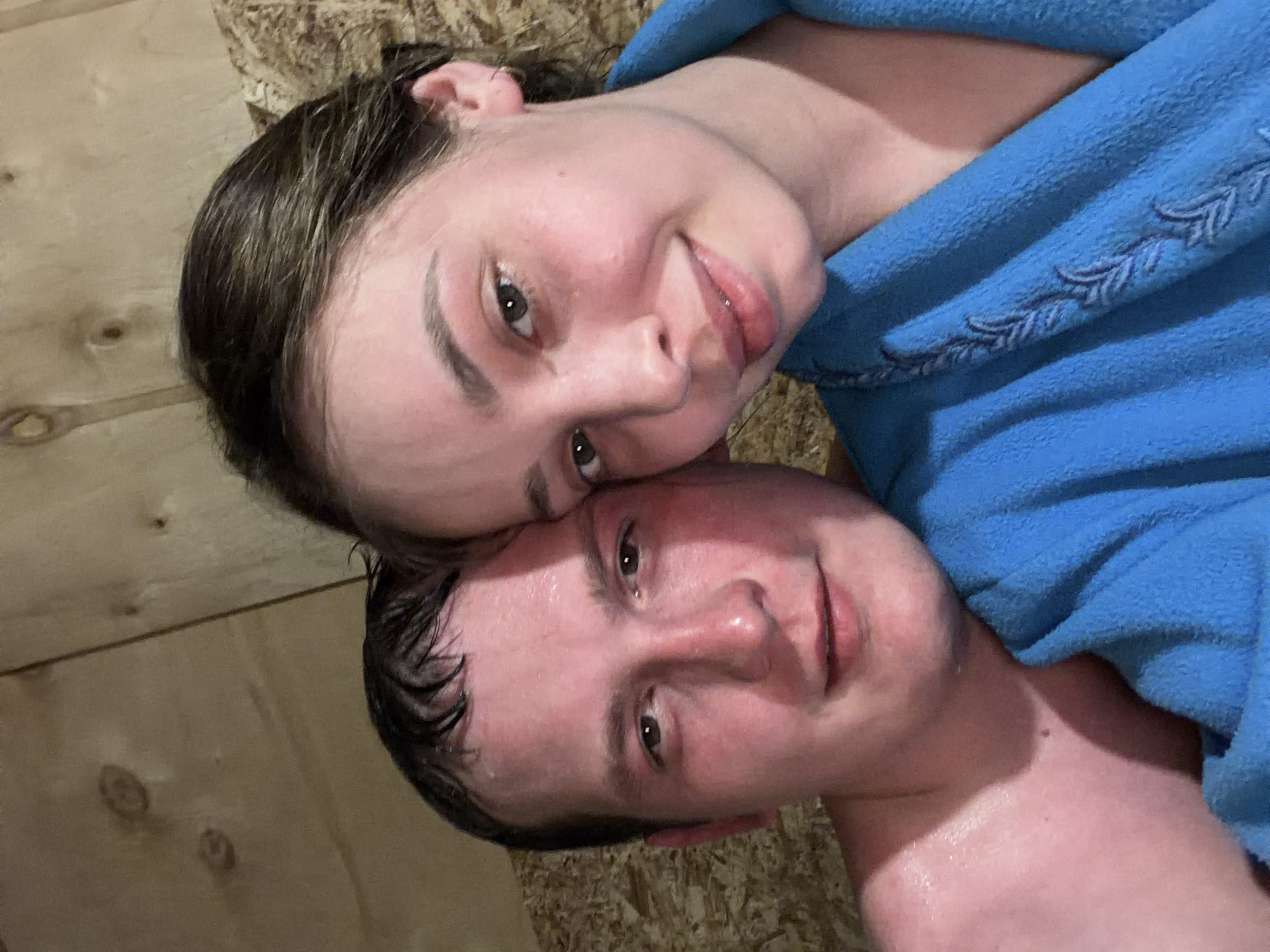 Two individuals are visible, a woman with damp dark brown hair lies above a man with wet, dark hair. The woman is smiling slightly, and her head rests on the man's. The man also appears to be smiling slightly. A blue bathrobe is partially visible, covering part of the woman. The background appears to be a wood-paneled wall or ceiling. Based on the prompt's inclusion of "Кемуль, Russia", it can be deduced that the location is likely within this city. The scene suggests they may be in a sauna or a bathroom setting.