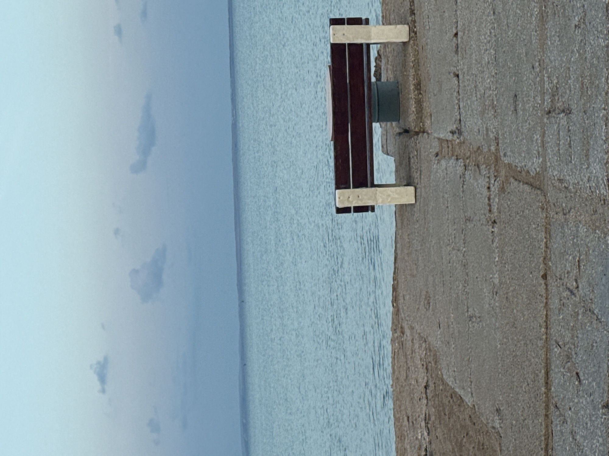 An empty wooden bench with beige legs is situated on a stone-paved embankment overlooking a large body of calm, light blue water. The sky is a pale blue with scattered, wispy clouds. The bench is positioned towards the right of the frame, facing the water. The embankment is made of large, rectangular stone slabs with visible cracks and wear. The water extends to the horizon, where a faint landmass is visible. The scene appears to be a serene, possibly coastal, setting.