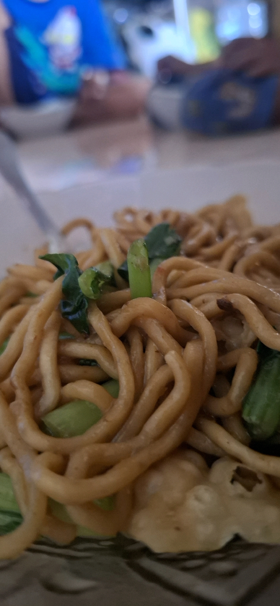A close-up, shallow depth-of-field shot focuses on a dish of noodles with green vegetables. The noodles are a golden brown, coiled and tangled together, with some pieces of green bok choy interspersed. A fork is visible on the left side of the frame, partially submerged in the noodles. In the blurred background, two individuals are partially visible, with one wearing a blue shirt. The setting appears to be an indoor dining area. Based on the provided information, this image was taken in Cibinong Hilir, Indonesia.