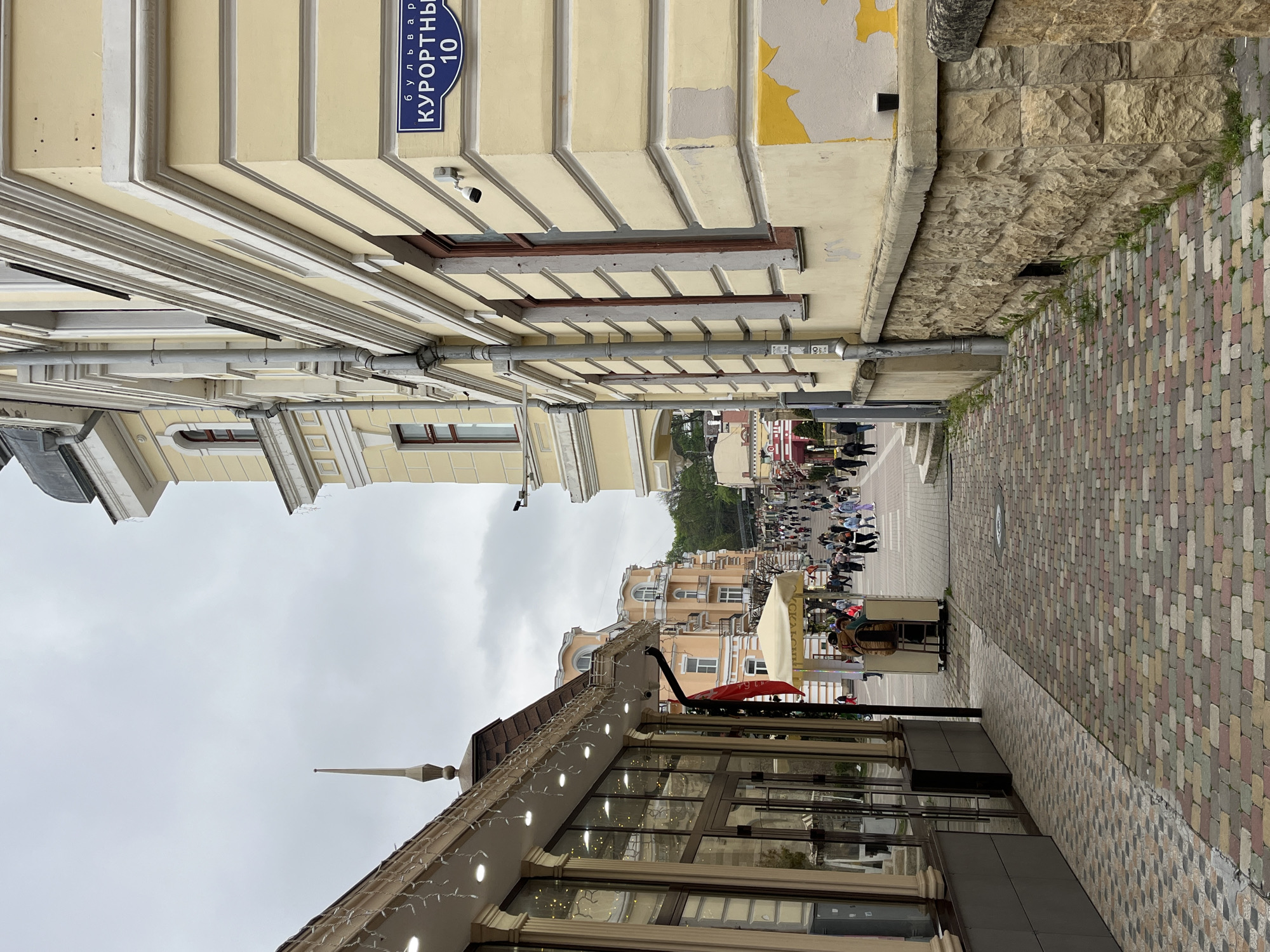 This outdoor shot shows a street in Kislovodsk, Russia. The view is slightly angled upwards, capturing the scene as if looking down a narrow passageway between buildings. Pavement brickwork lines the street on the right, while the architecture on either side creates a sense of enclosure. The street is populated with pedestrians in the distance, indicating a bustling urban environment. An establishment with a sign in Russian (readable as "Куррортный 10", meaning "Resort 10") is visible on a nearby building, indicating a commercial establishment. A restaurant is seen on the lower part of the image, it has its windows closed. The weather appears overcast, and the overall ambiance suggests a vibrant yet confined urban space.